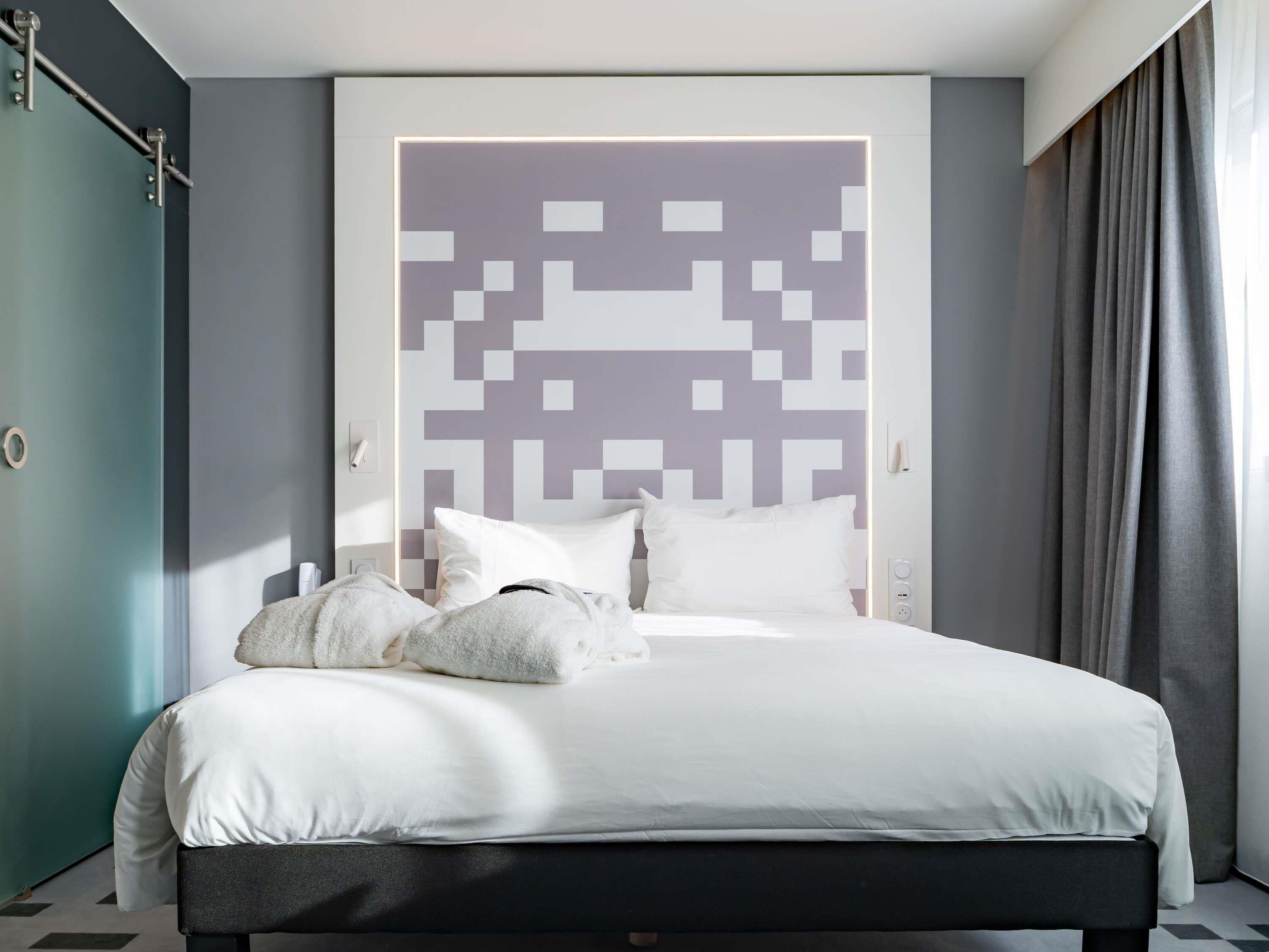 Une chambre d’hôtel moderne, équipée d’un lit, d’une tête de lit ornée d’un motif géométrique, d’une armoire et d’une fenêtre avec des rideaux.