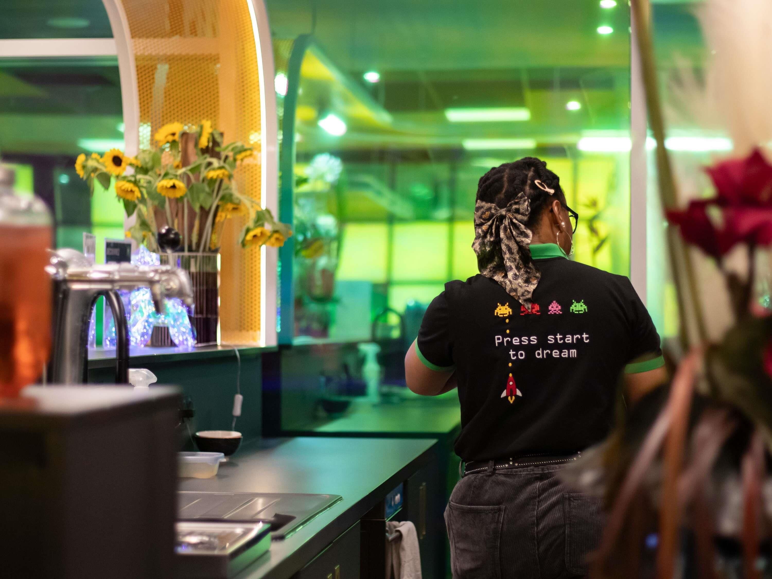 Una persona con una camiseta negra que lleva un mensaje en la espalda, parada en una habitación con iluminación verde y un mostrador con varios objetos.