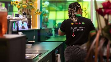 Una persona con una camiseta negra que lleva un mensaje en la espalda, parada en una habitación con iluminación verde y un mostrador con varios objetos.