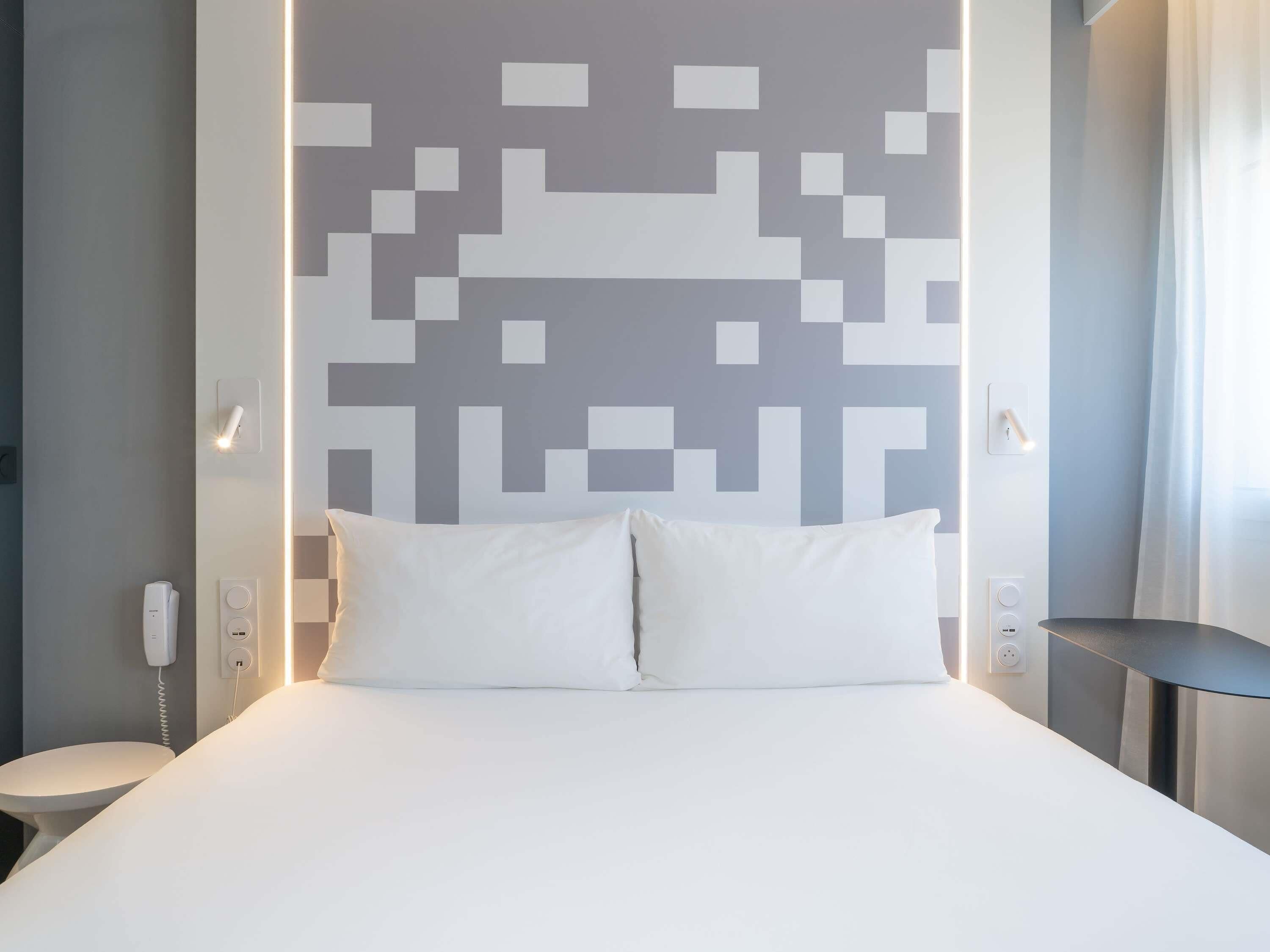 A modern hotel room with a bed, a bedside table, a wall-mounted light, and a wall with a geometric pattern.