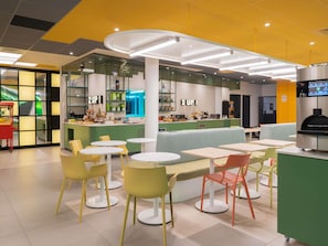 Un café moderno con mesas redondas, sillas de colores, una barra con menú y un horno de pizza.