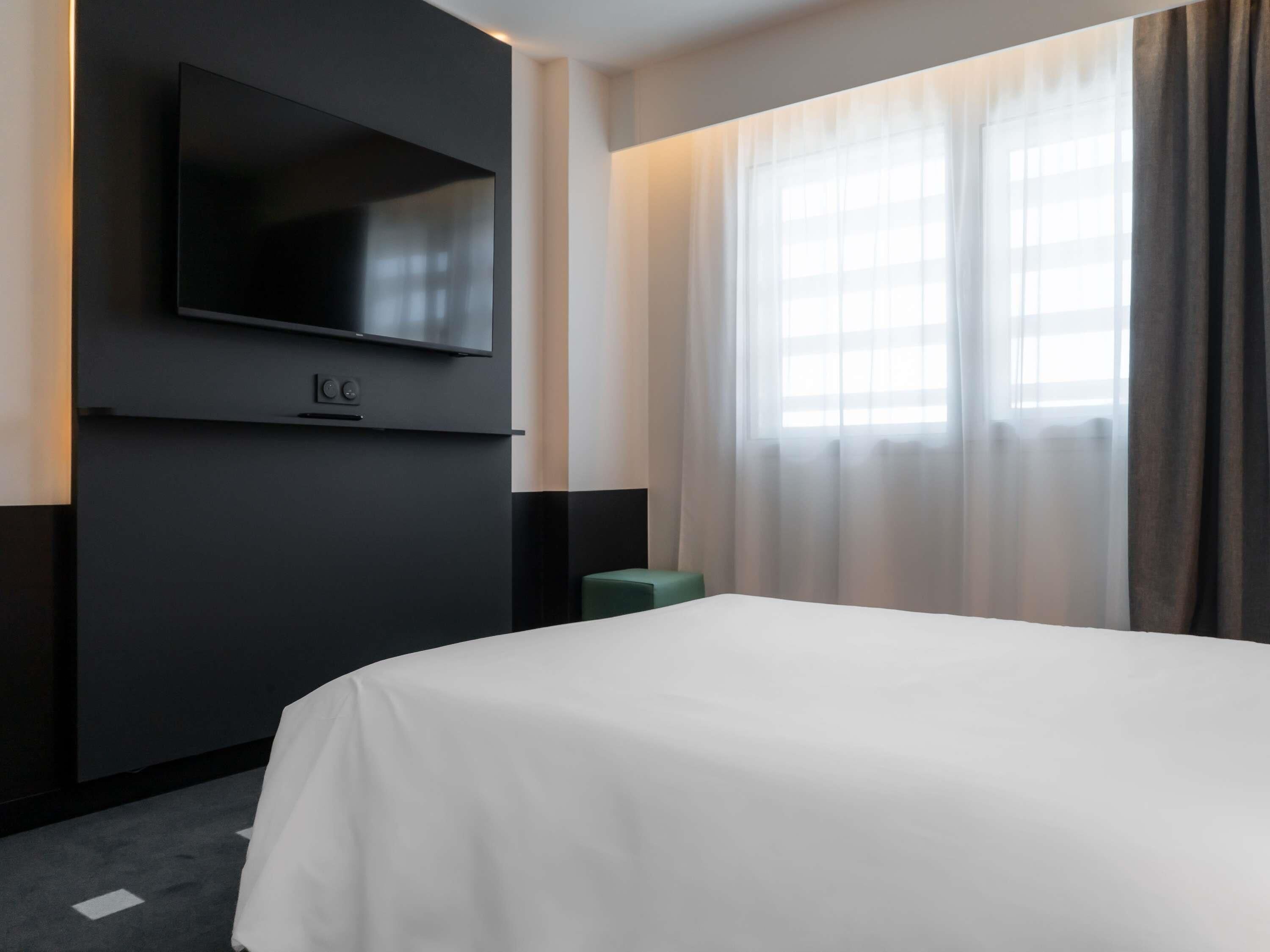 A hotel room with a large bed, a flat-screen TV mounted on the wall, and a window with sheer curtains.