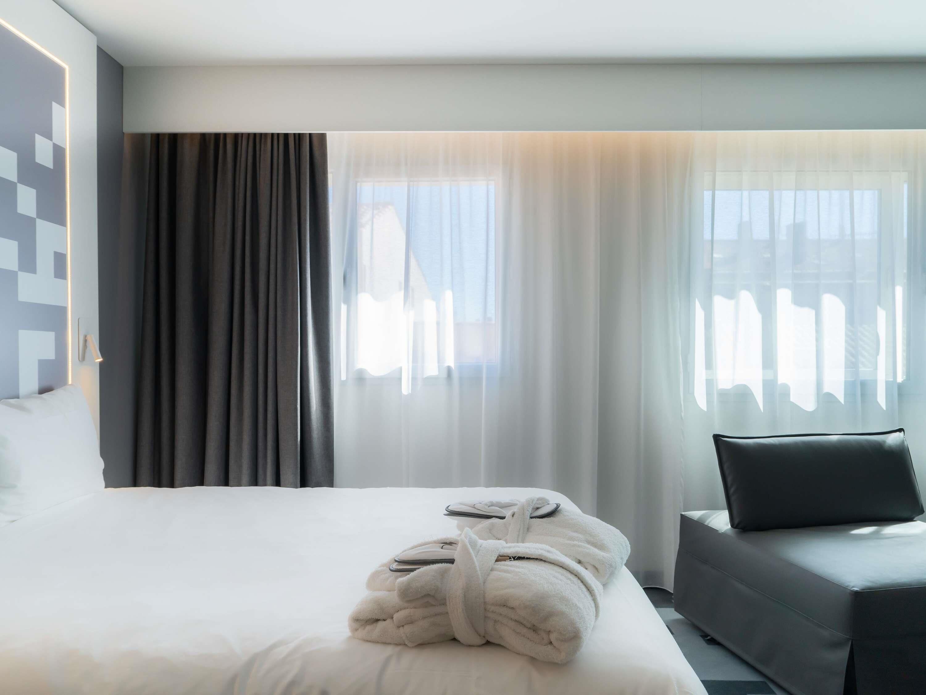 Une chambre d’hôtel avec un lit soigneusement fait, une pile de serviettes pliées, un canapé noir et des rideaux légers laissant passer la lumière naturelle.