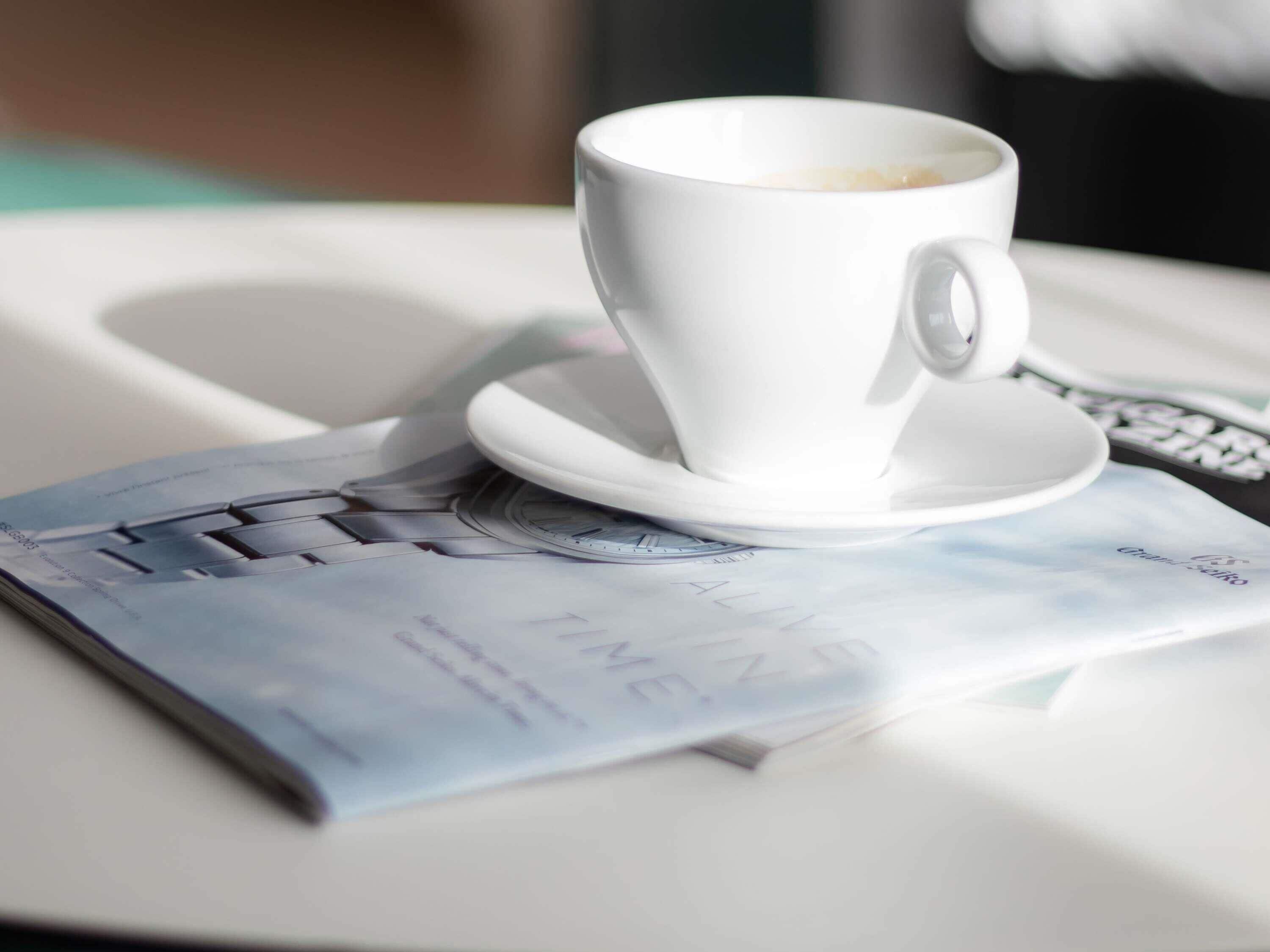 Une tasse de café blanche sur une table blanche, avec un magazine dont la couverture est bleue.
