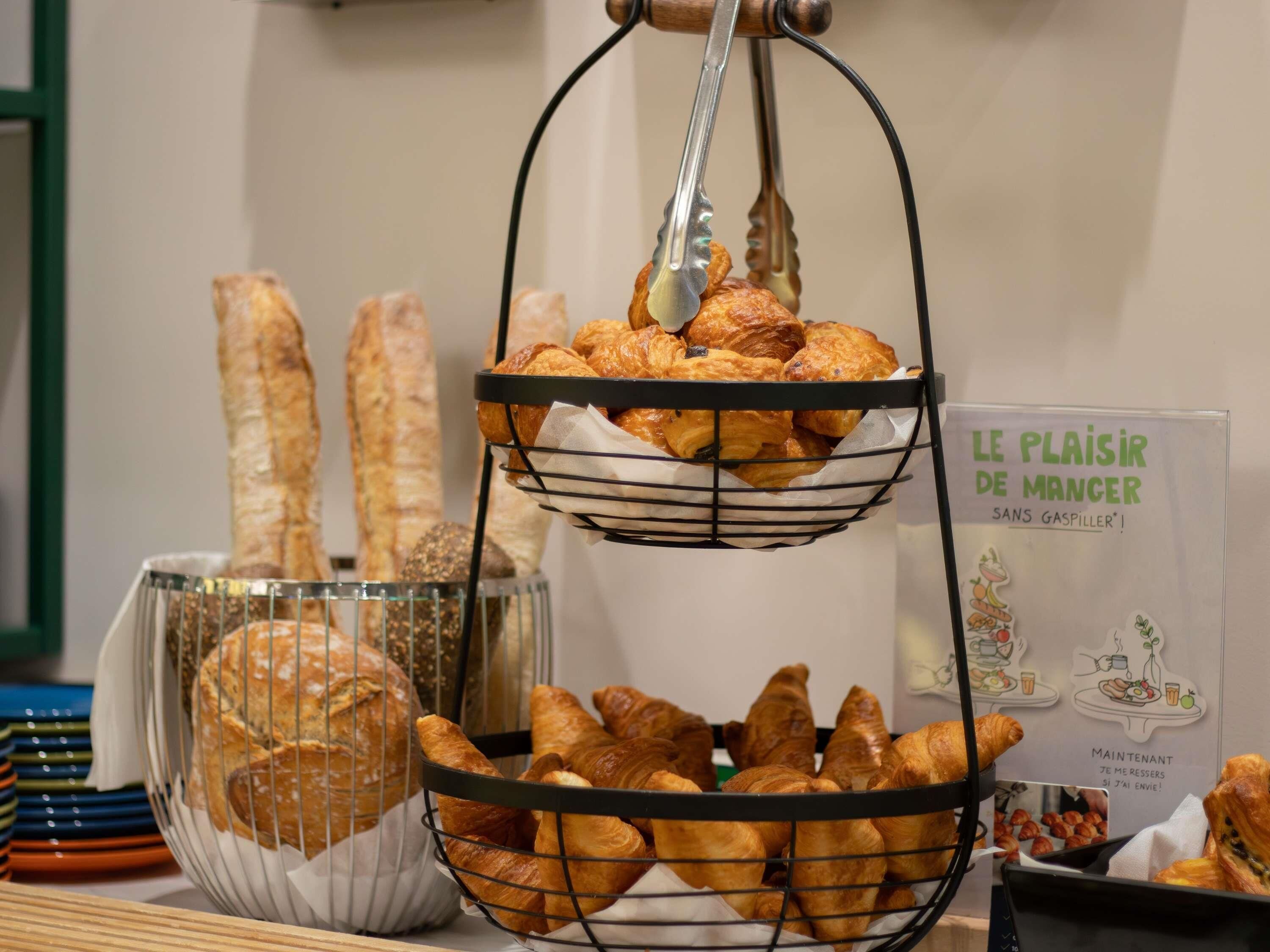 Una exhibición de pan y pasteles, incluyendo cruasanes y barras de pan, en una cesta y una cesta de alambre, con un letrero al fondo.