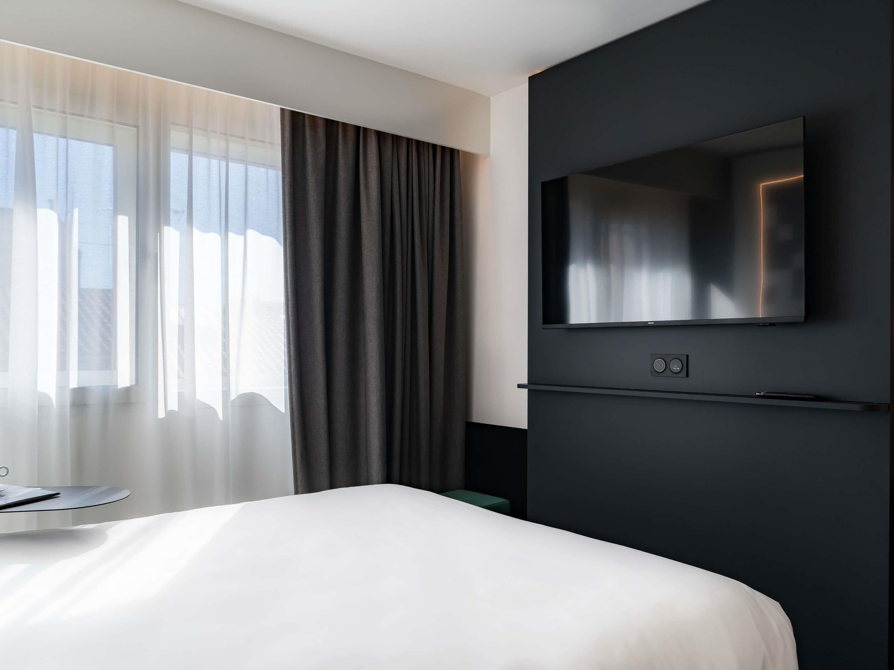 Habitación de hotel moderna con una cama, un televisor de pantalla plana montado en la pared y un ventanal con cortinas translúcidas.