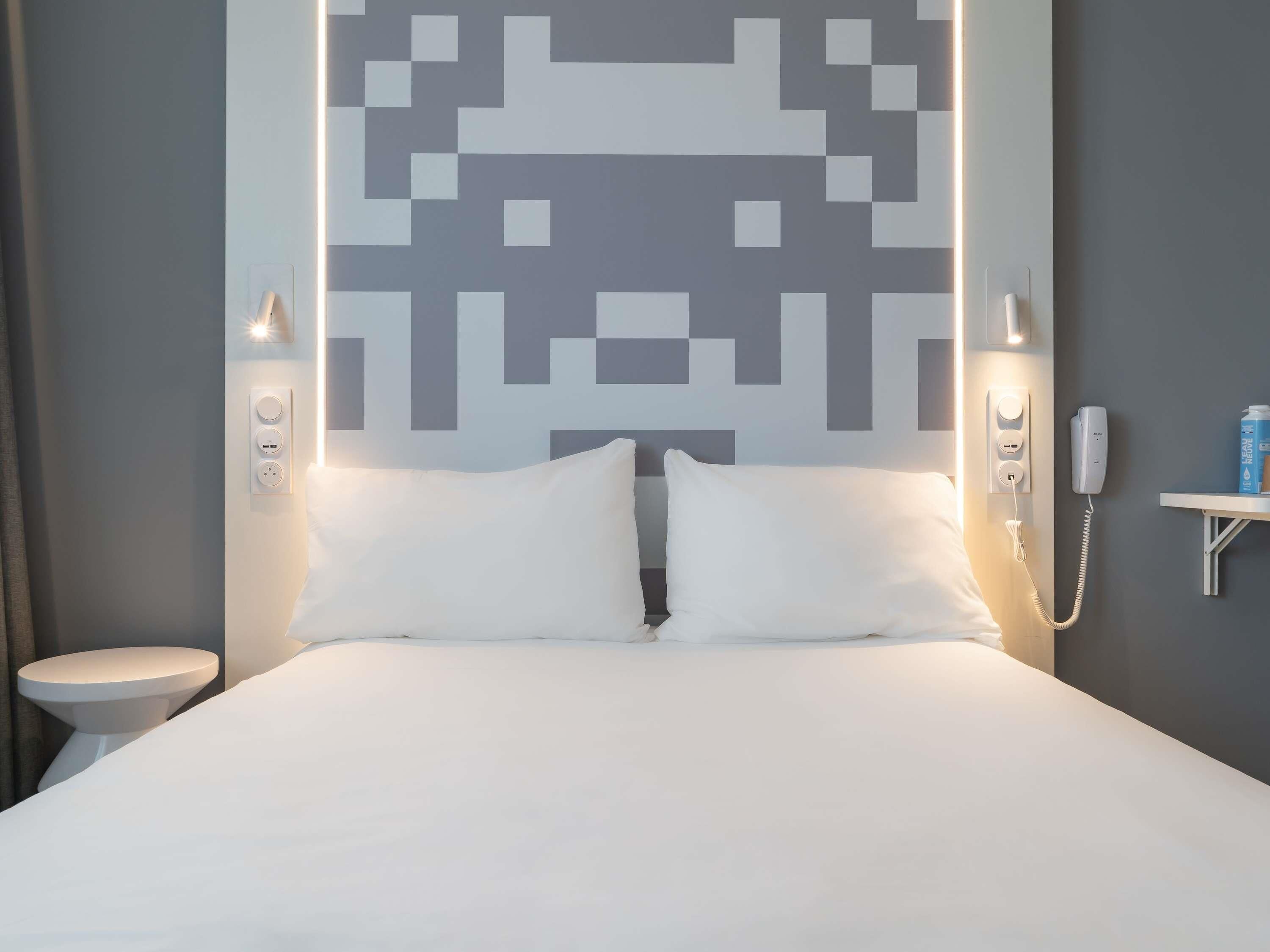 A modern hotel room with a large bed, white bedding, a wall-mounted phone, and a small bedside table with a bottle.