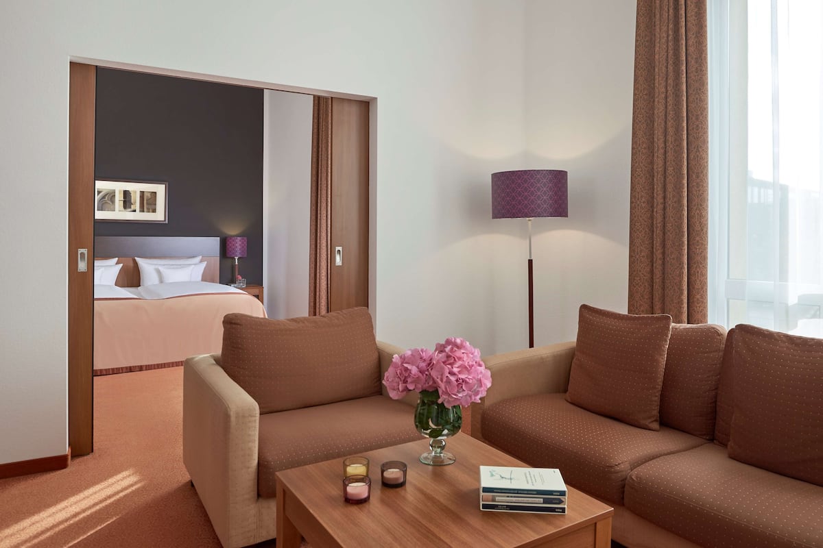 Habitación de hotel con sofá, una mesa de centro con un florero, una lámpara y una cama con mesita de noche.