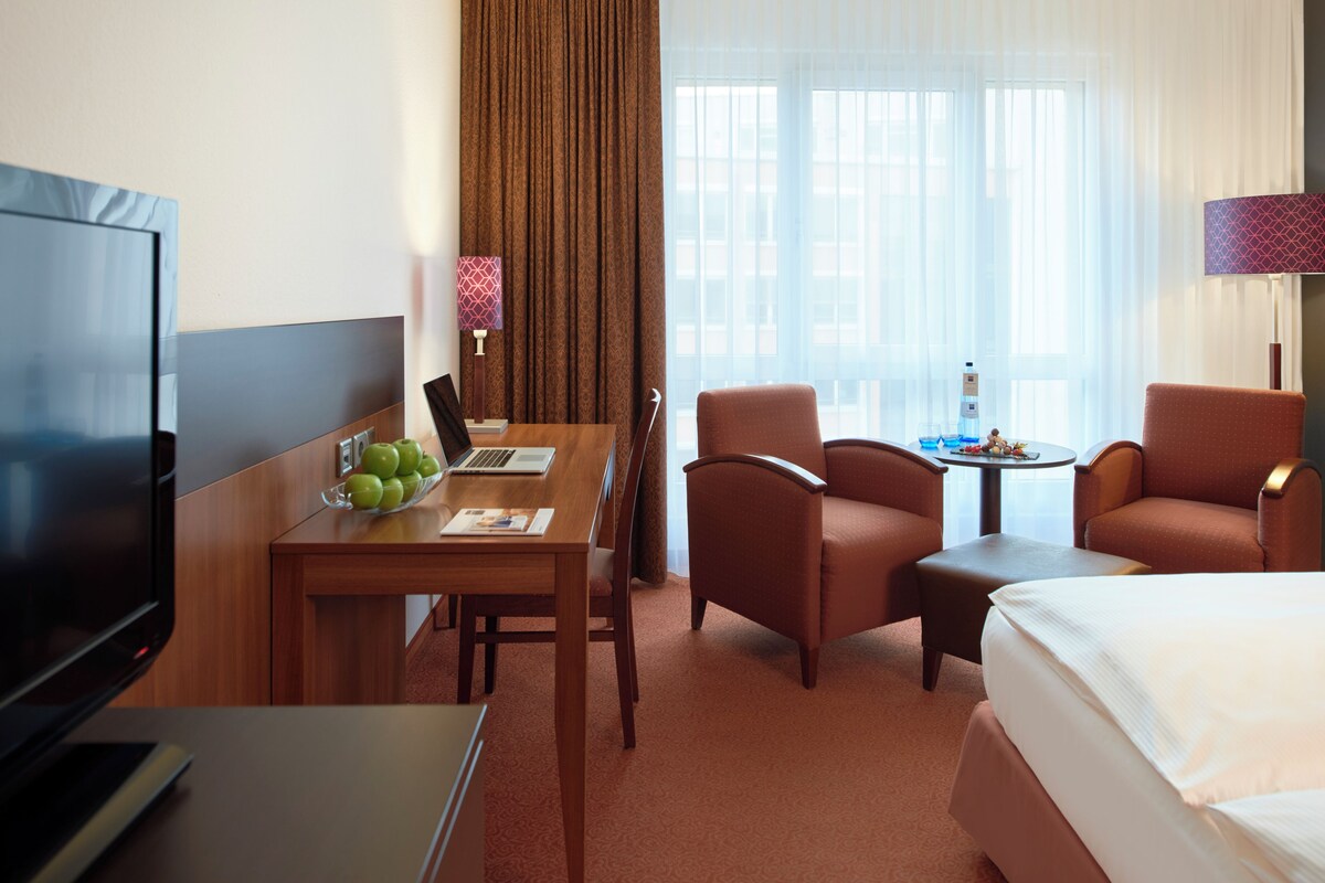 A hotel room with a bed, a desk with a laptop, a television, two armchairs, a small table with a bottle and fruit, and a window with curtains.