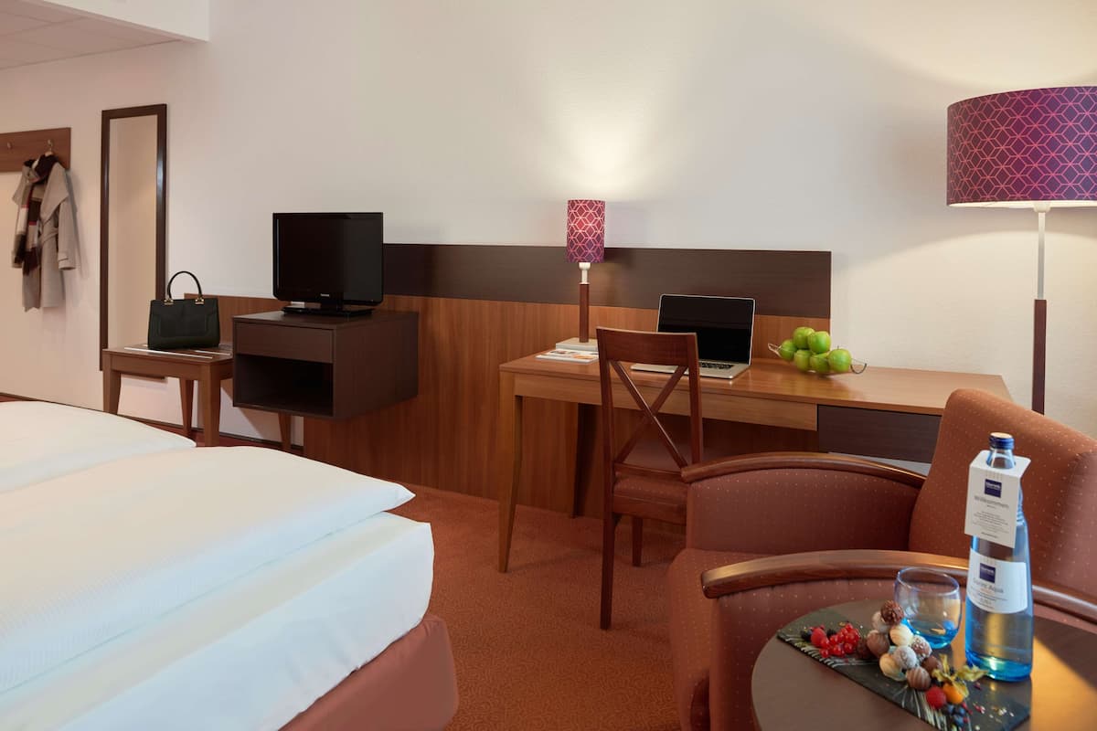 Habitación de hotel con cama, escritorio con una computadora portátil, televisor, silla, mesa con aperitivos y lámpara.