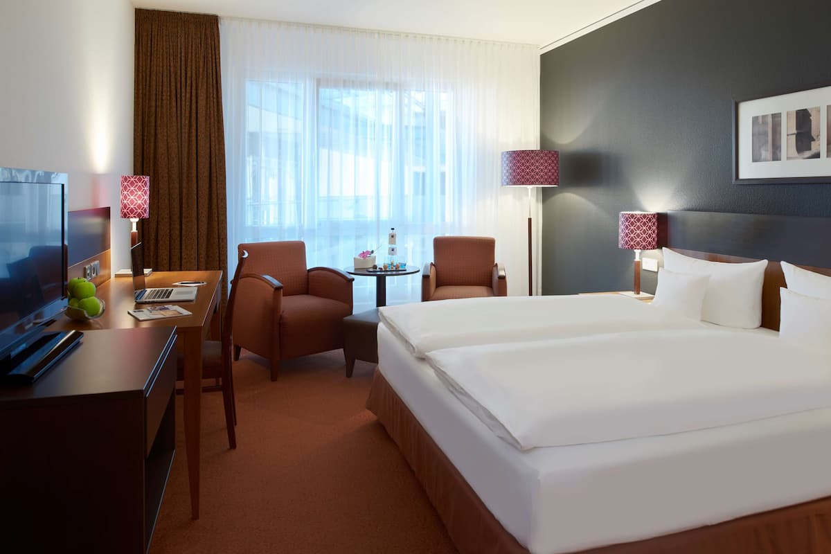 Habitación de hotel con una cama grande, dos sillones, un escritorio con una computadora portátil, un televisor y una pequeña mesa con un objeto decorativo.