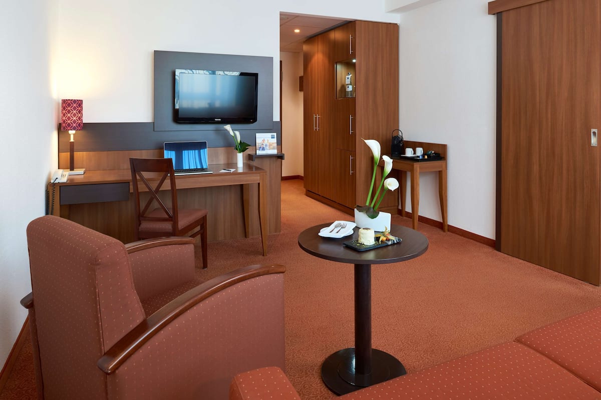 Una habitación de hotel con zona de estar, una mesita con un florero, un televisor montado en la pared y un armario de madera.