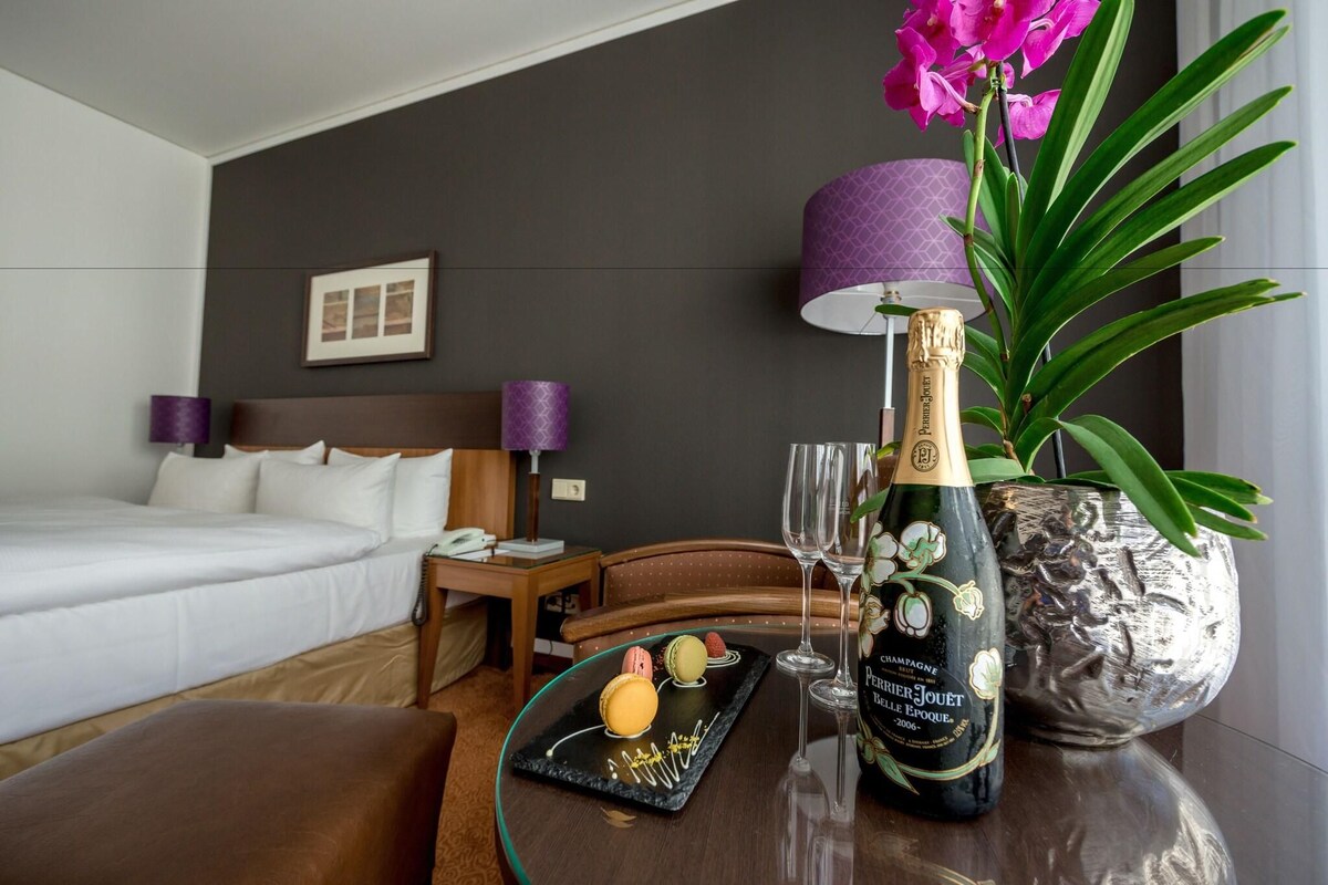 A hotel room with a bed, a bedside table with a lamp, a decorative vase with flowers, a bottle of champagne, and a tray with macarons.