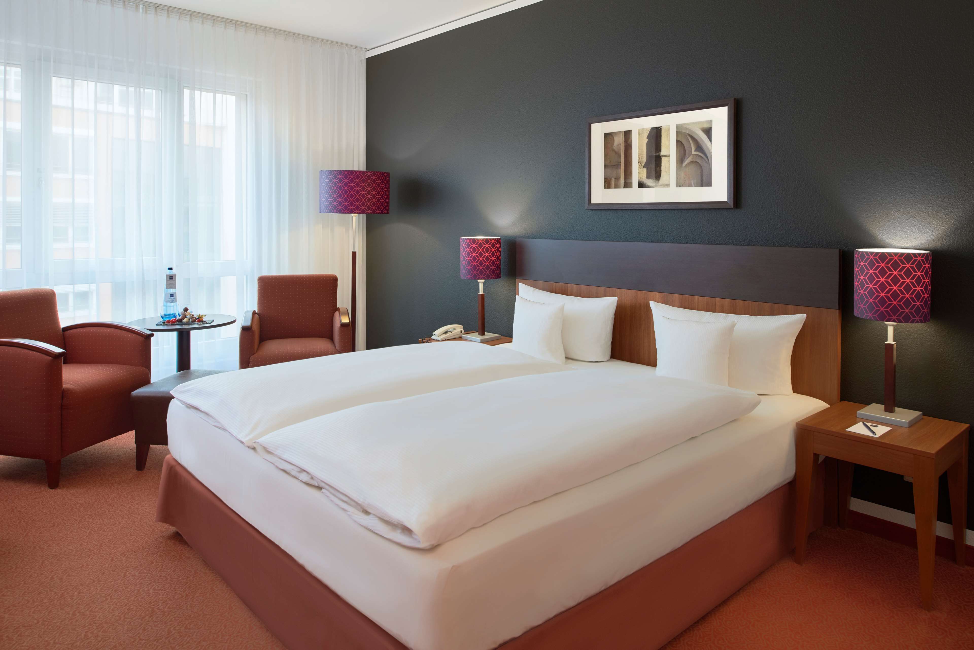 A hotel room with a large bed, two bedside tables, a chair, a desk, and a window with sheer curtains.