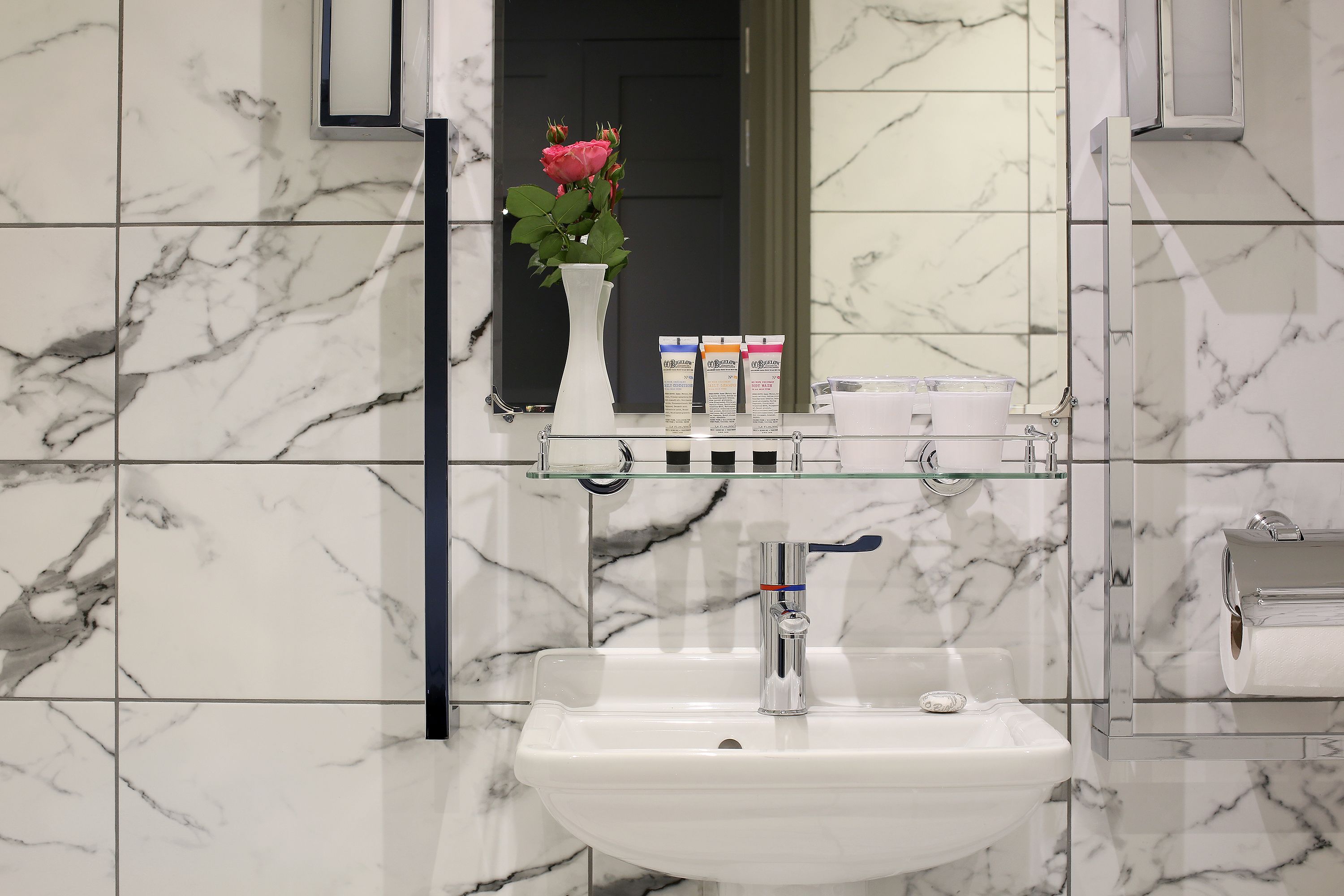 Un baño con lavamanos de mármol blanco, repisa de vidrio con artículos de aseo y un florero con flores.