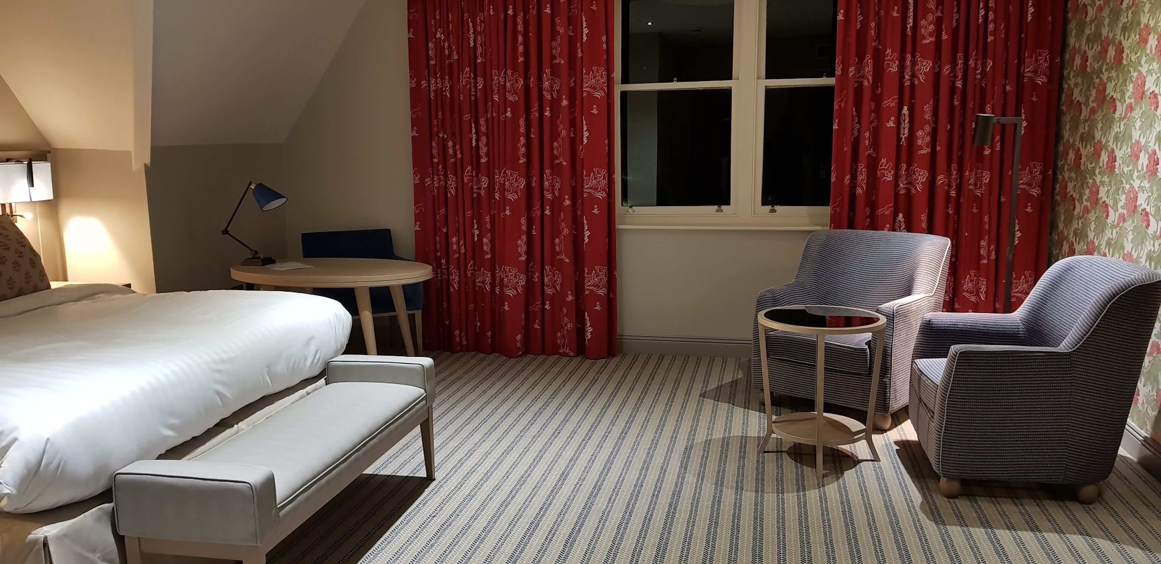 Une chambre d’hôtel avec un lit, un bureau, deux fauteuils et une fenêtre avec des rideaux rouges.