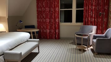 Una habitación de hotel con una cama, un escritorio, dos sillones y una ventana con cortinas rojas.