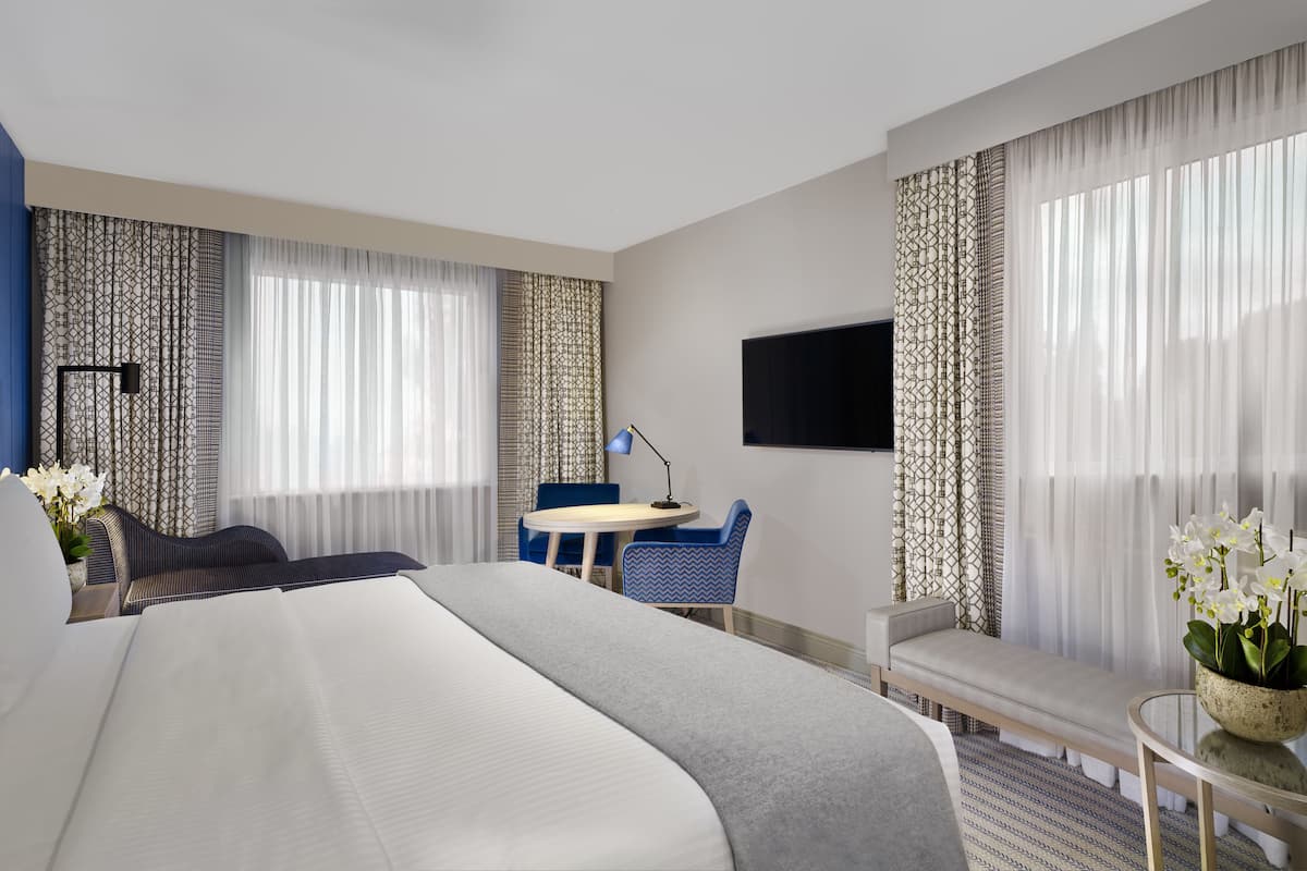 Ein Hotelzimmer mit einem großen Bett, einem Schreibtisch mit Stuhl, einem an der Wand befestigten Fernseher und einem kleinen Tisch mit Pflanze.