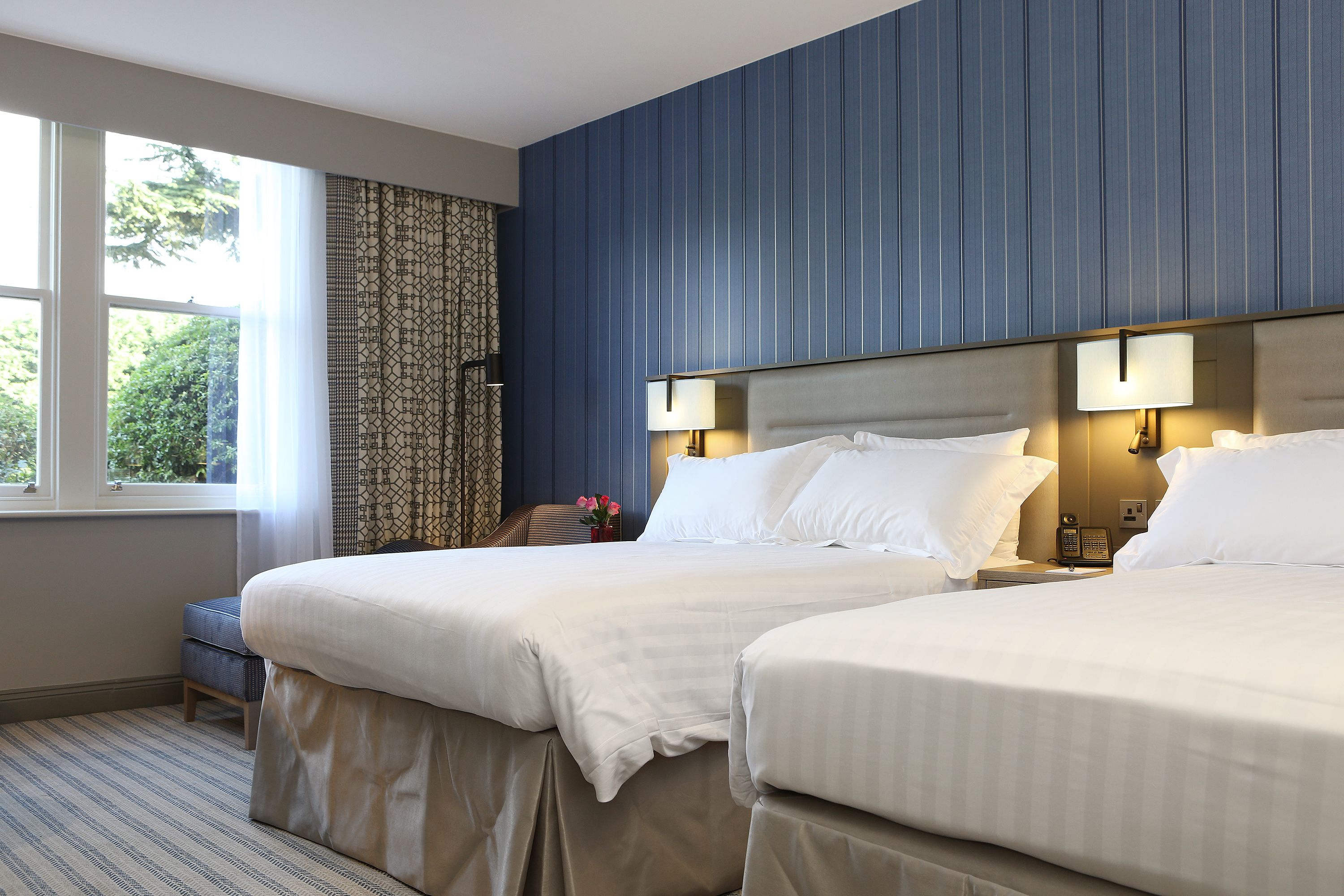 A hotel room with two beds, a window with curtains, a bedside table with a lamp, and a small seating area with a chair and a vase of flowers.