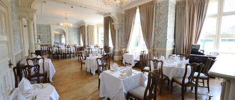 A spacious dining hall with multiple tables set for dining, elegant chandeliers, and large windows with curtains.