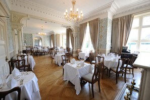 Une salle à manger spacieuse, avec de nombreuses tables dressées, de magnifiques lustres et de grandes fenêtres ornées de rideaux.