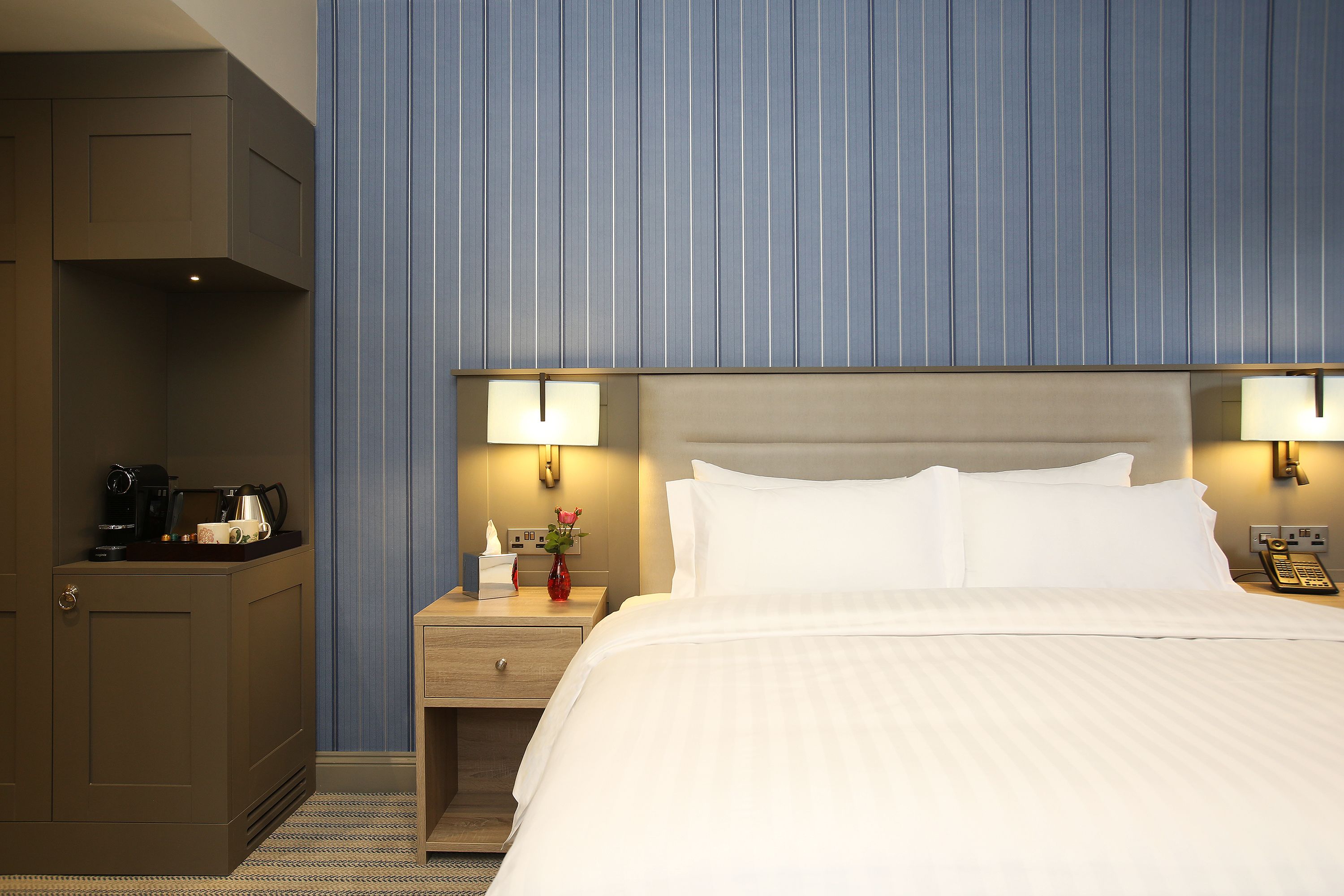 A hotel room with a bed, bedside lamps, a nightstand with a phone, a vase with flowers, and a coffee maker.