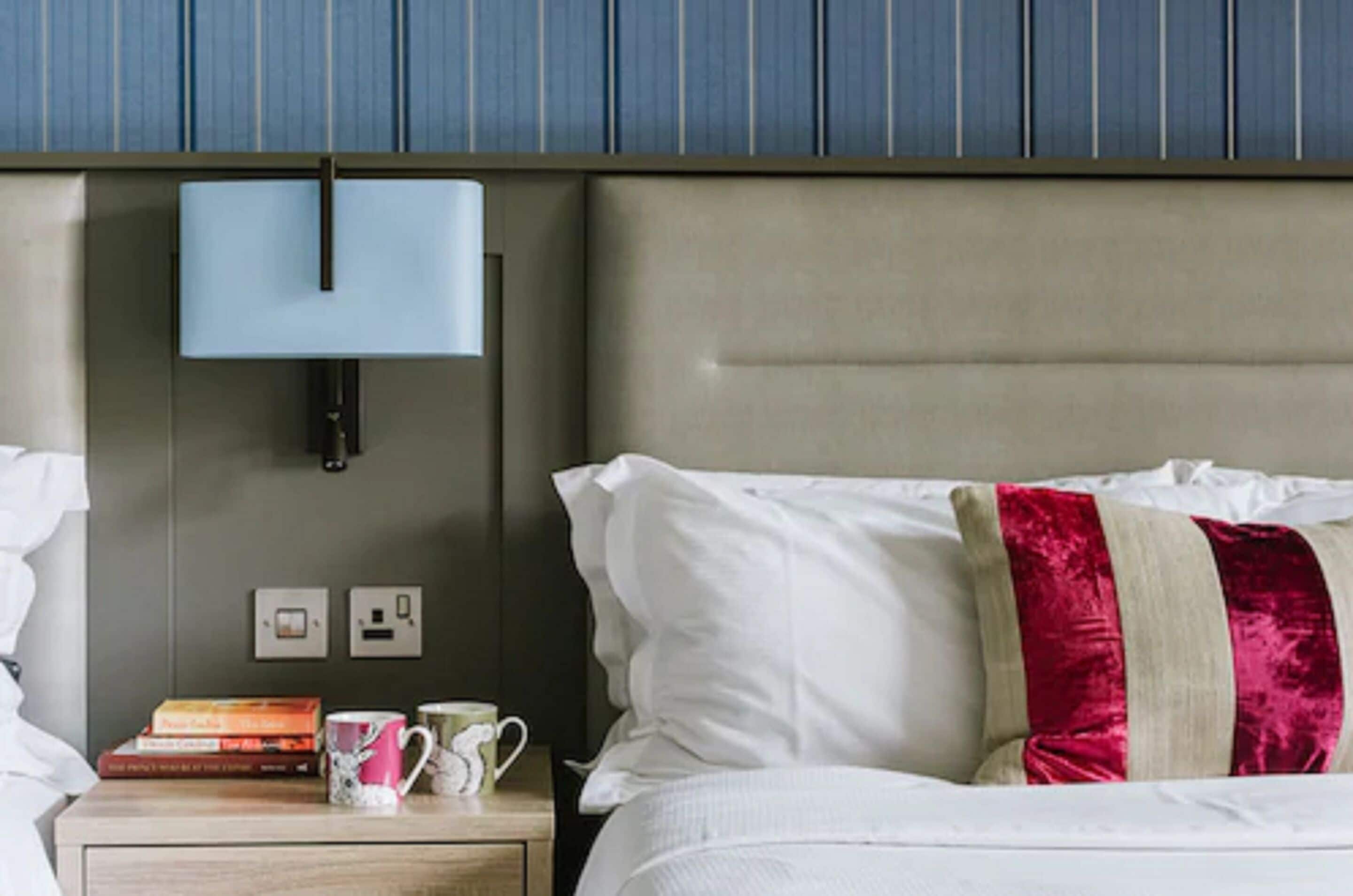 Een opgemaakt bed met witte en rode kussens, een nachtkastje met boeken en een mok, en een wandlamp.