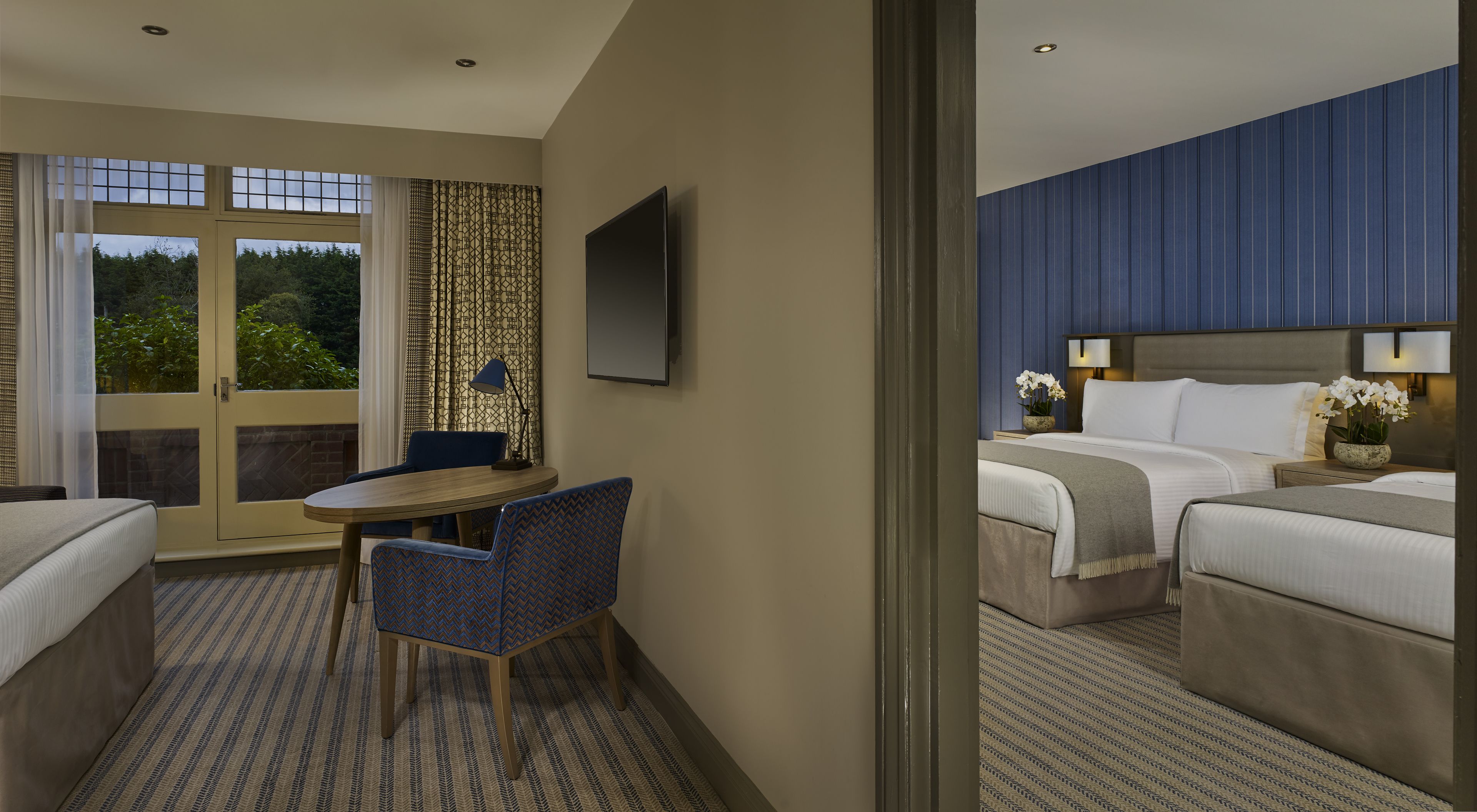 A hotel room with a bed, a desk with a chair, a TV, and a view of greenery through the window.