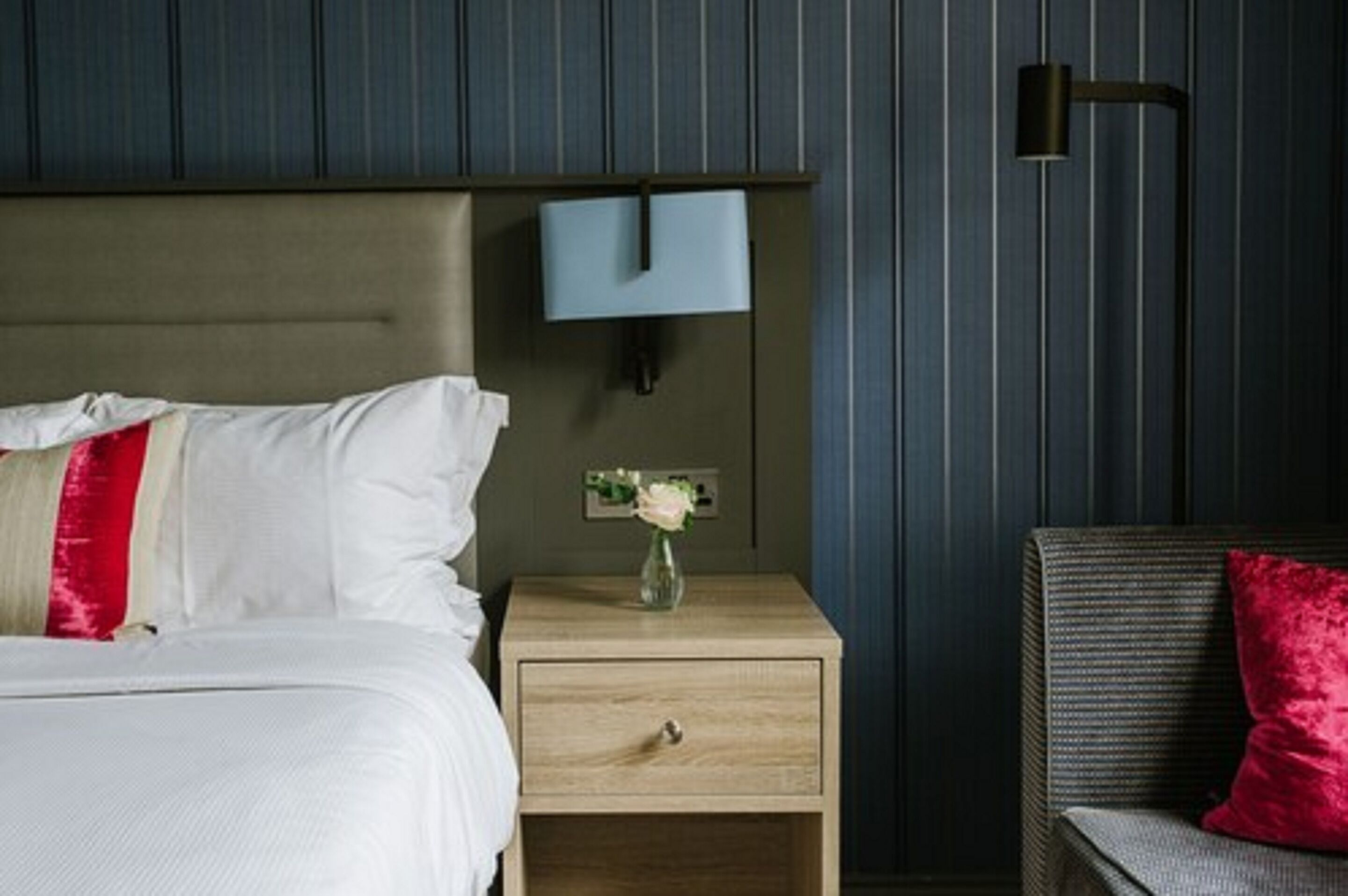 A neatly made bed with white and red pillows, a bedside table with a vase of flowers, and a wall-mounted light fixture.