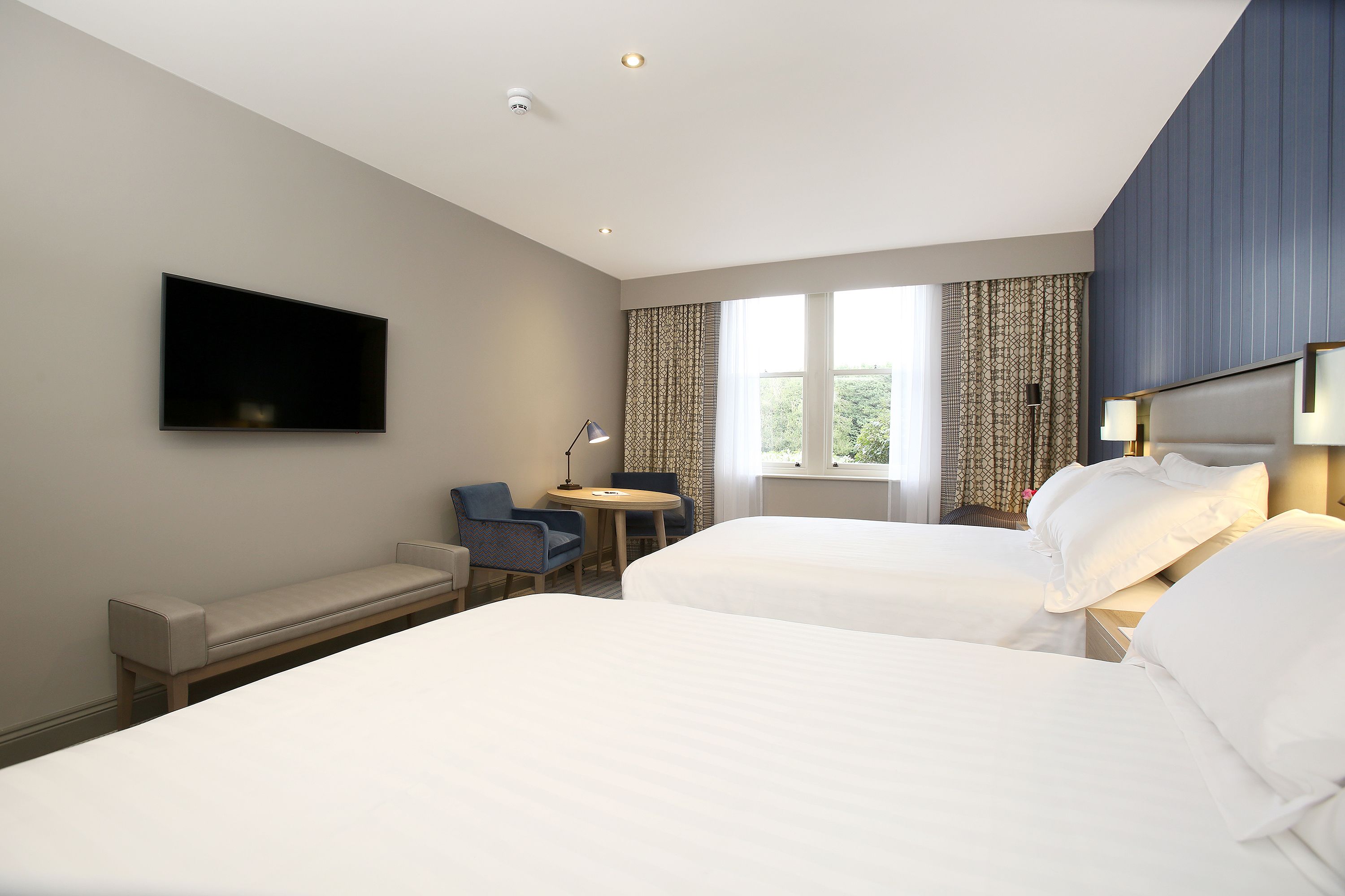 A hotel room with two beds, a desk, a chair, a TV, and a window with curtains.