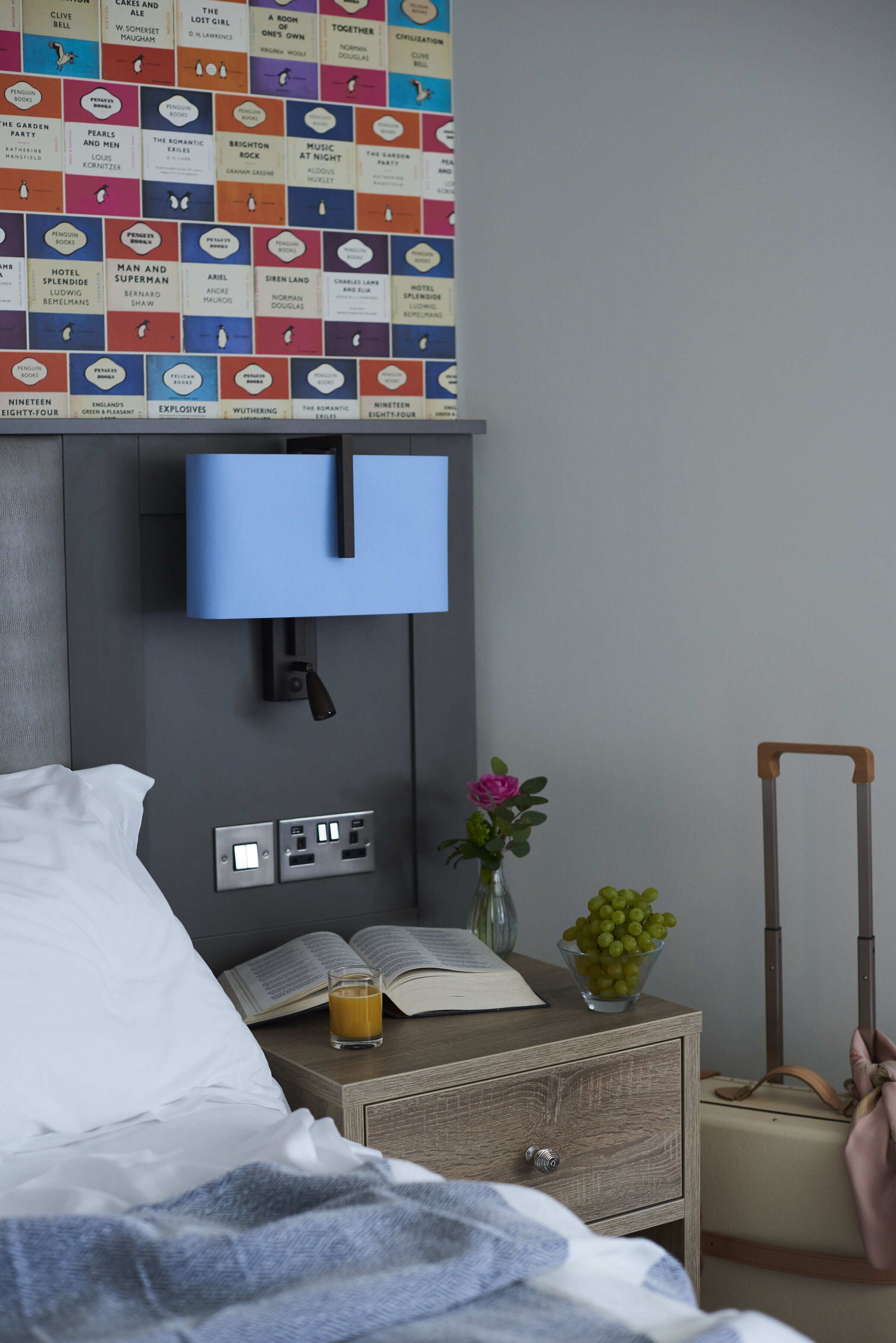 Ein Hotelzimmer mit Bett, Nachttisch mit Buch, Glas Saft, Blumenvase und Koffer.