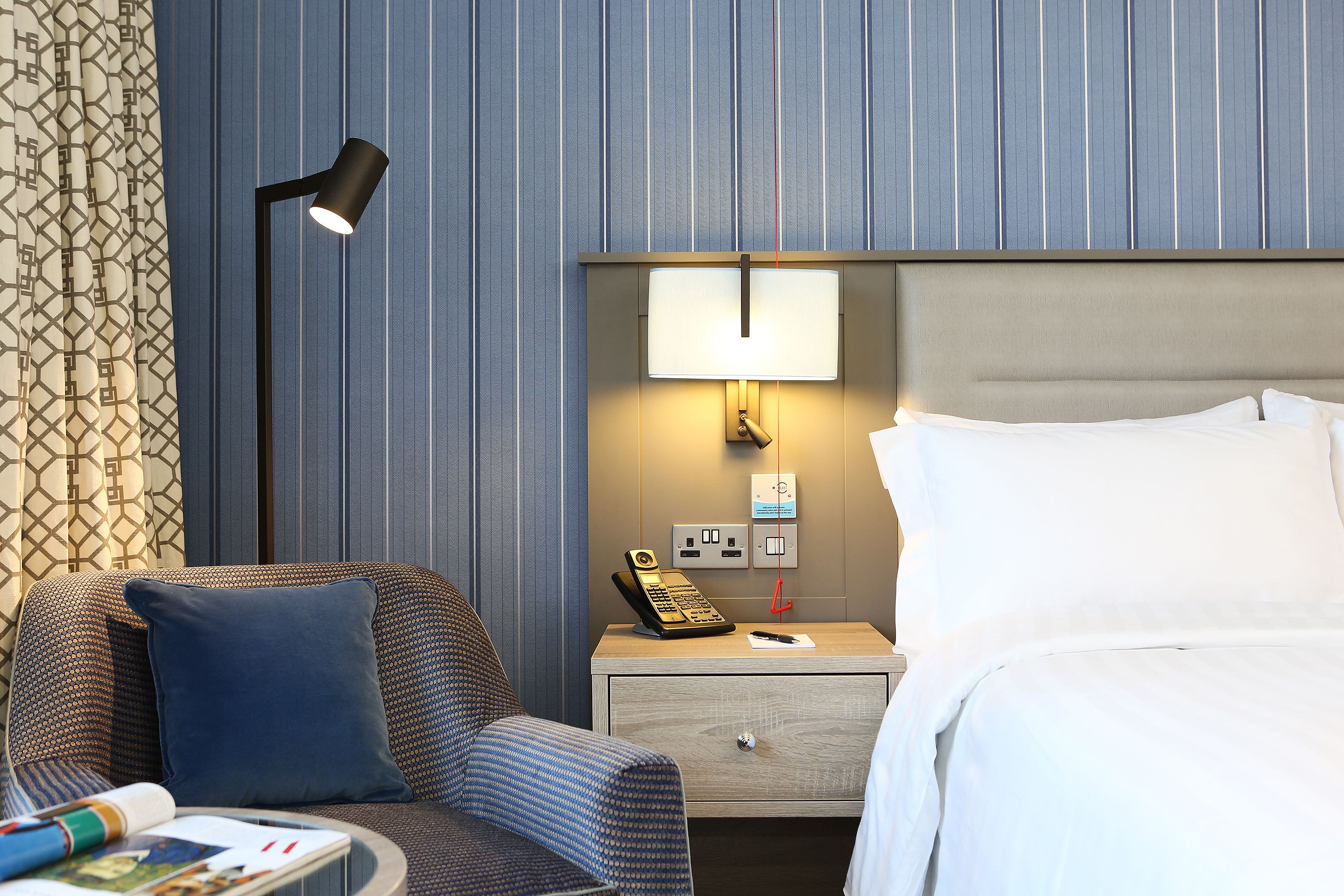 A hotel room with a bed, a nightstand with a lamp, a telephone, and a wall-mounted light.