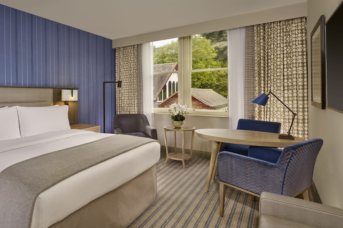 Ein Hotelzimmer mit Bett, Nachttisch, Lampe, Stuhl, kleinem Tisch mit Vase und Blick auf ein Gebäude durch das Fenster.