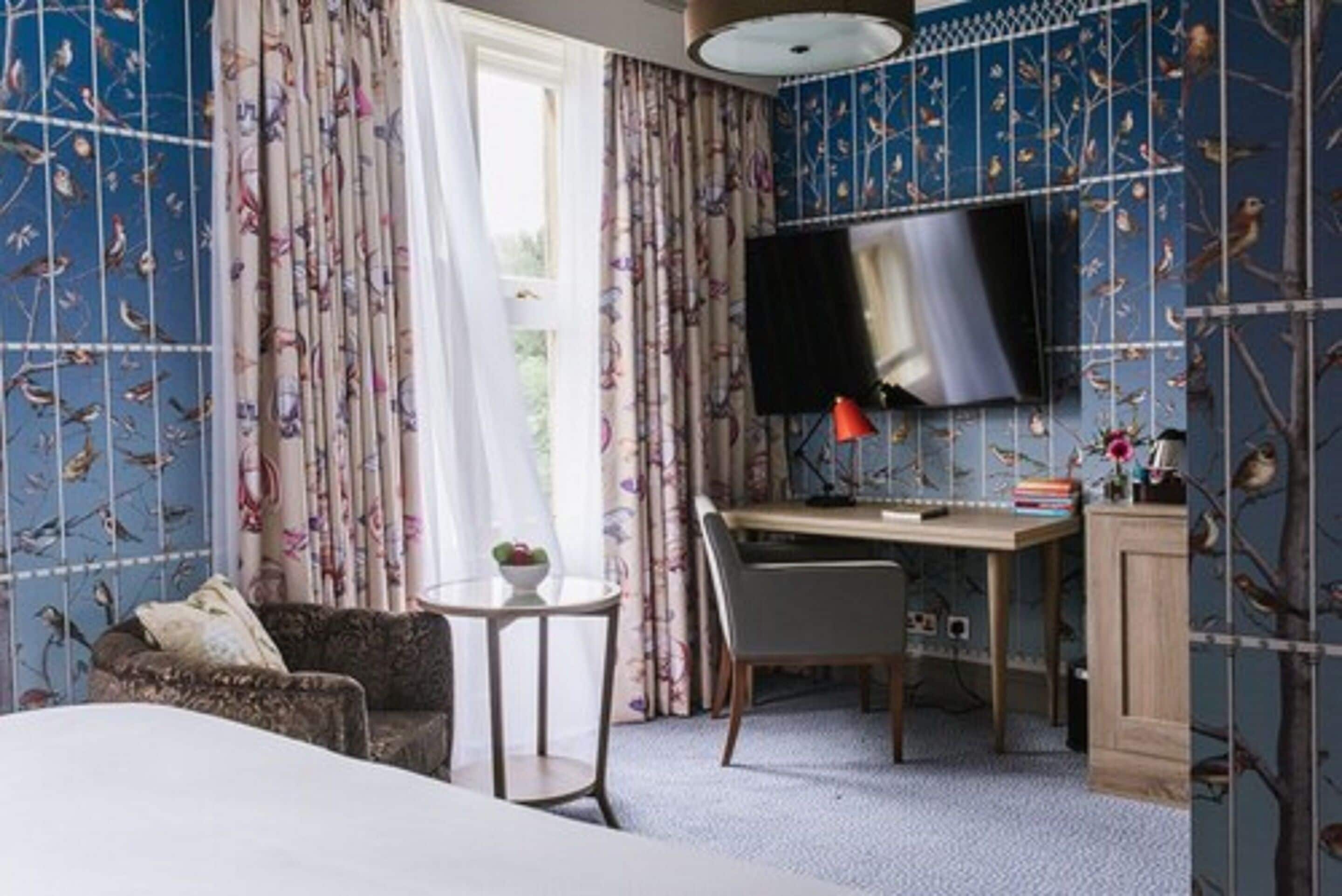 A bedroom with a bed, a desk, a chair, a TV, and curtains with a bird pattern.