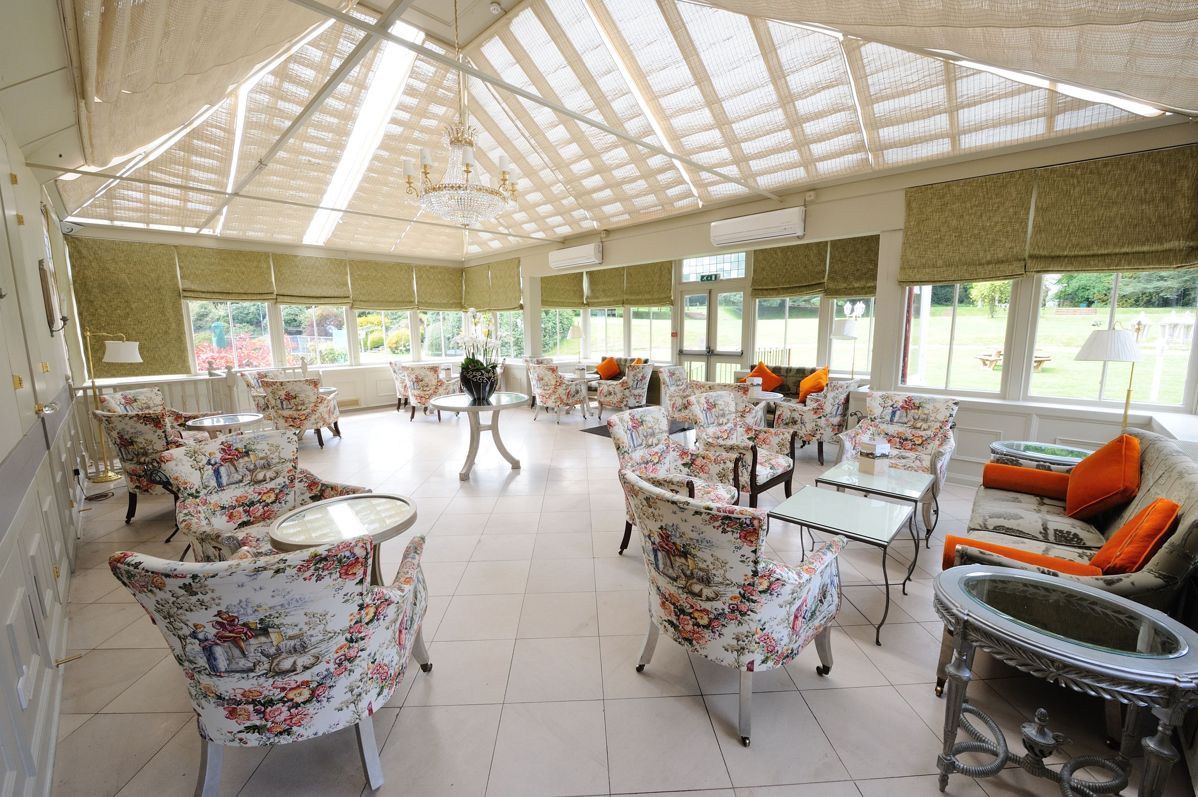 Een ruime binnenruimte met stoelen en tafels met bloemenprint, grote ramen en een hoge plafondconstructie van glas.