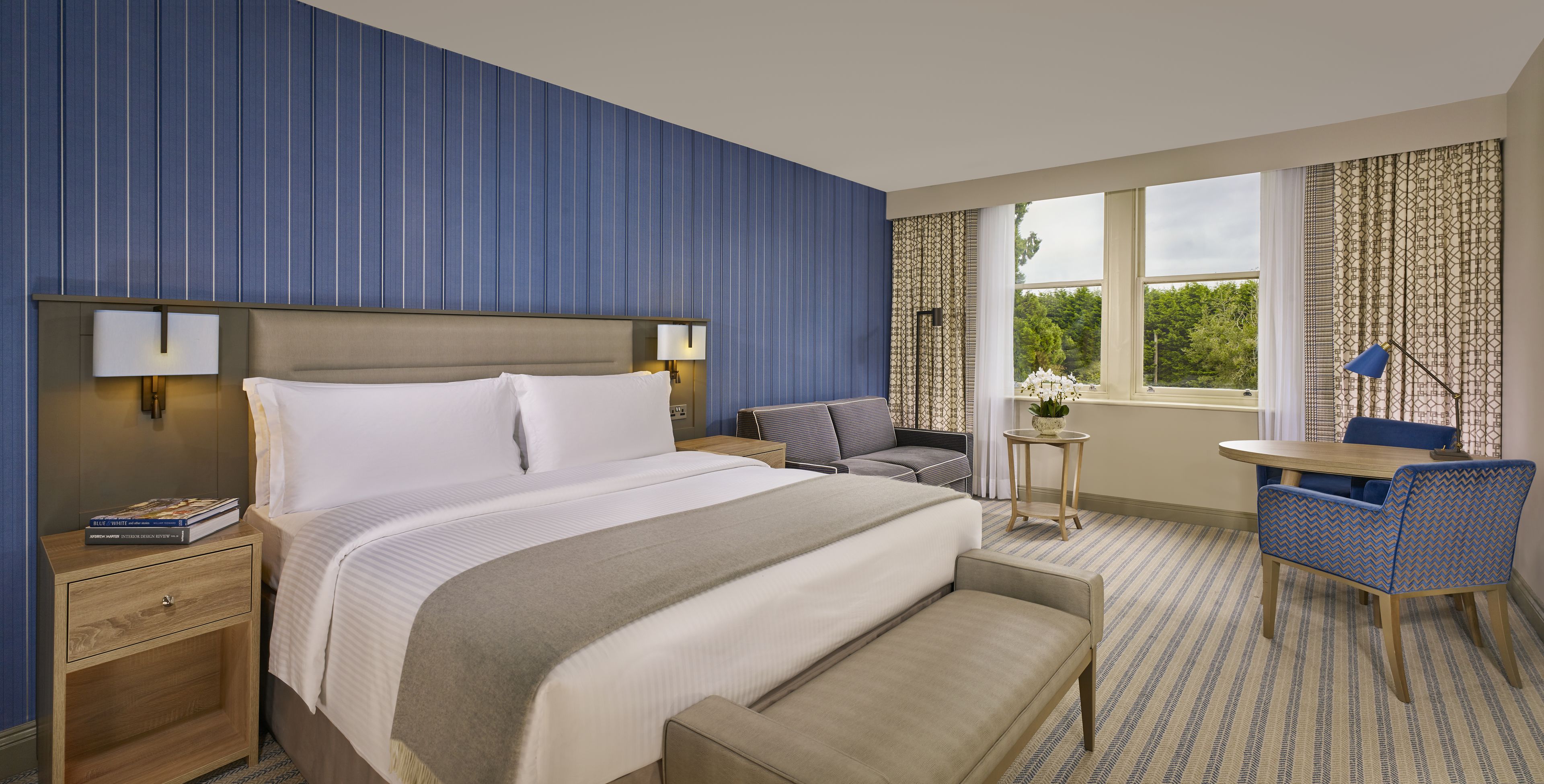 Une chambre d’hôtel avec un grand lit, des tables de chevet, une petite table avec une lampe, une chaise et une vue sur l’extérieur.