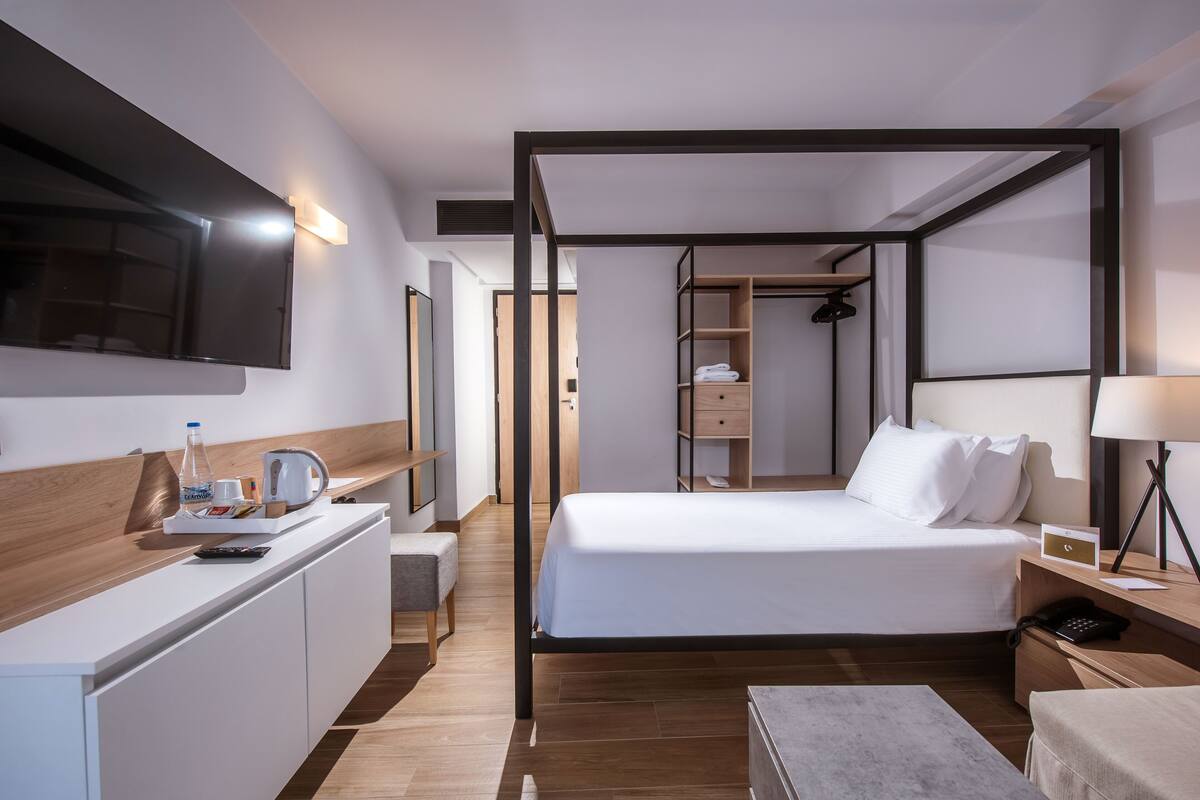 A modern hotel room with a large bed, a desk with a flat-screen TV, a bedside table with a lamp, and a walk-in closet.