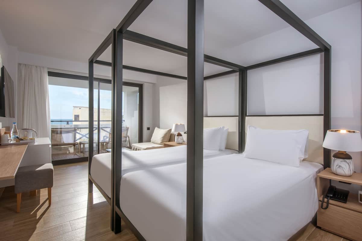 A modern hotel room with a four-poster bed, a desk with a chair, a small table, and a balcony with a view of the sea.
