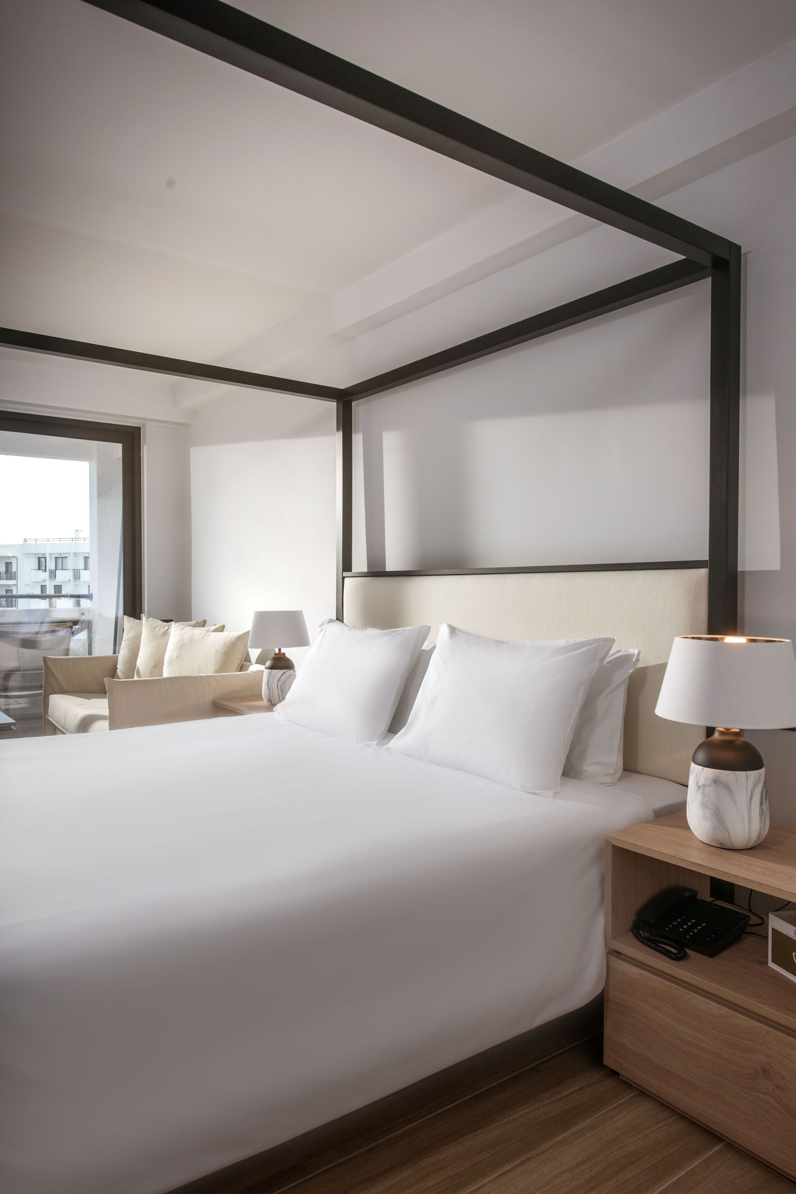 A modern hotel room with a large bed, a bedside table with a lamp, and a view of the city through the window.
