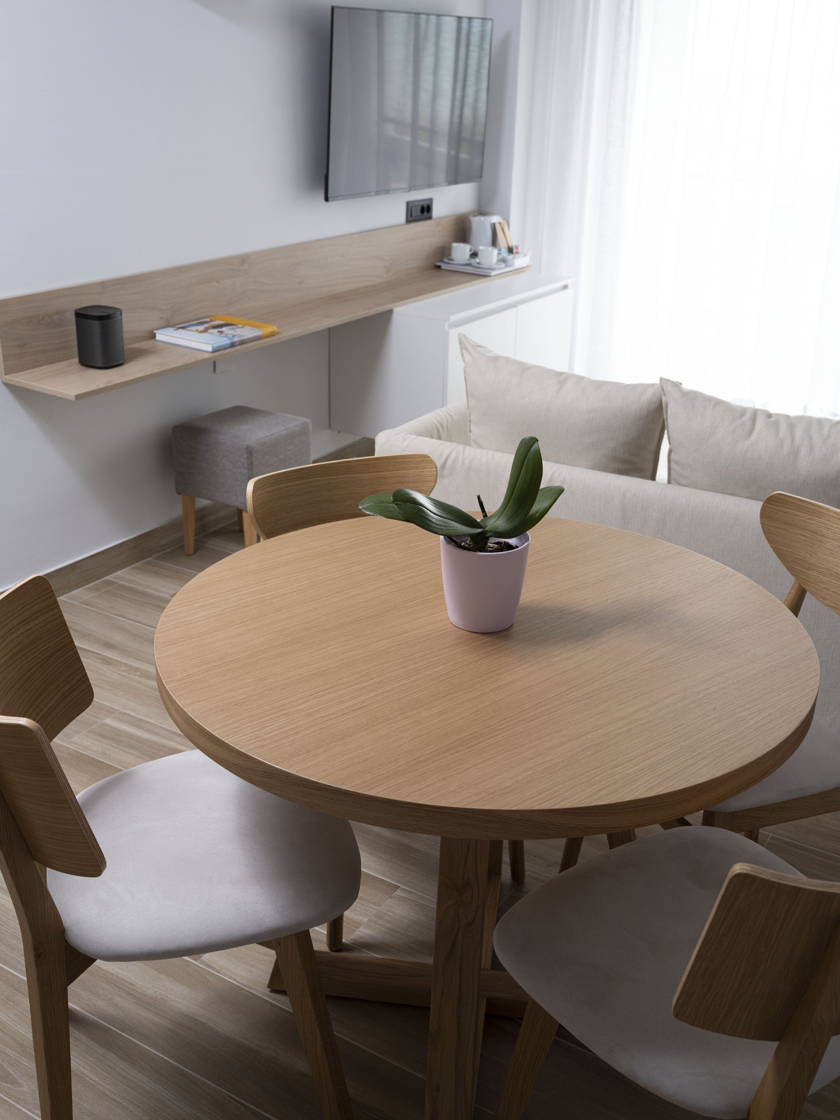 A modern dining area with a round wooden table, four chairs, a small potted plant, a sofa, and a flat-screen TV mounted on the wall.