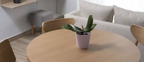 A modern dining area with a round wooden table, four chairs, a small potted plant, a sofa, and a flat-screen TV mounted on the wall.