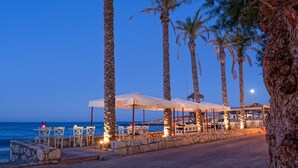 A seaside restaurant with outdoor seating, palm trees, and a view of the ocean. - Hersonissos Palace - All Inclusive (Hersonissos)