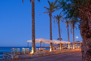 A seaside restaurant with outdoor seating, palm trees, and a view of the ocean. - Hersonissos Palace - All Inclusive (Hersonissos)
