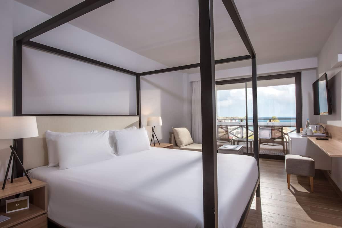 A modern hotel room with a large bed, a desk, a chair, and a balcony with a view of the sea.
