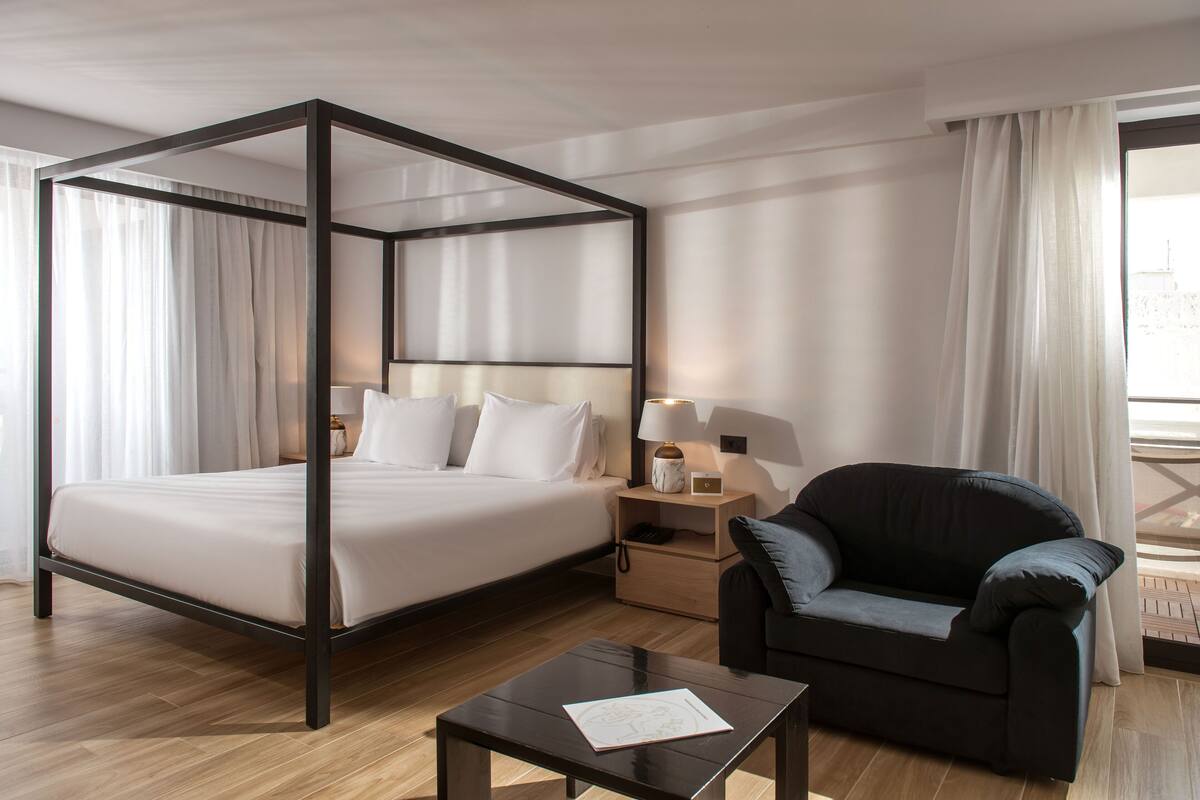 A four-poster bed with white linens, a dark-colored armchair, a bedside table with a lamp, and a wooden floor.