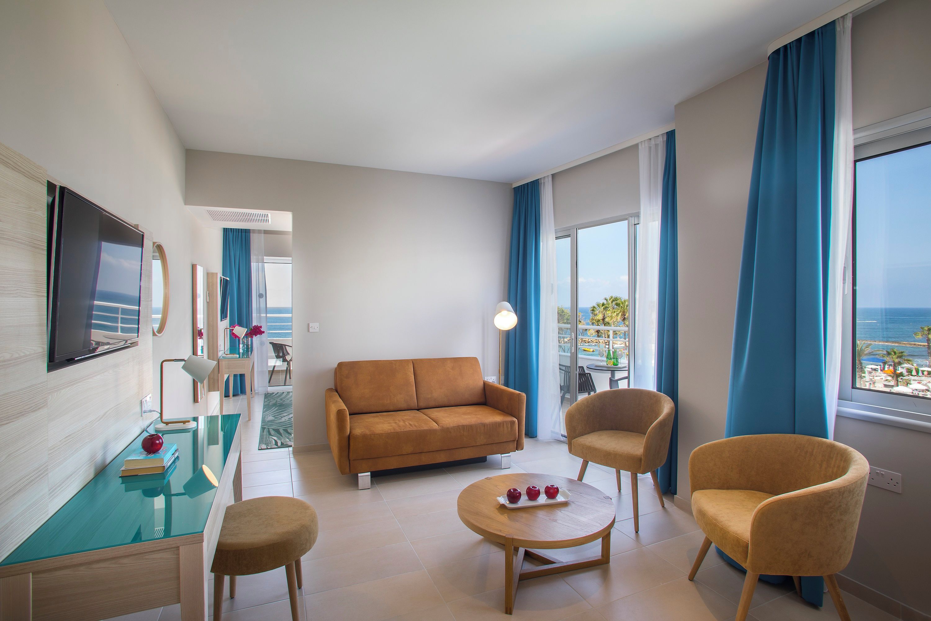 Una habitación de hotel moderna con sofá, sillas, una mesita con manzanas, televisor y vista al mar.