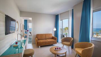 Una habitación de hotel moderna con sofá, sillas, una mesita con manzanas, televisor y vista al mar.