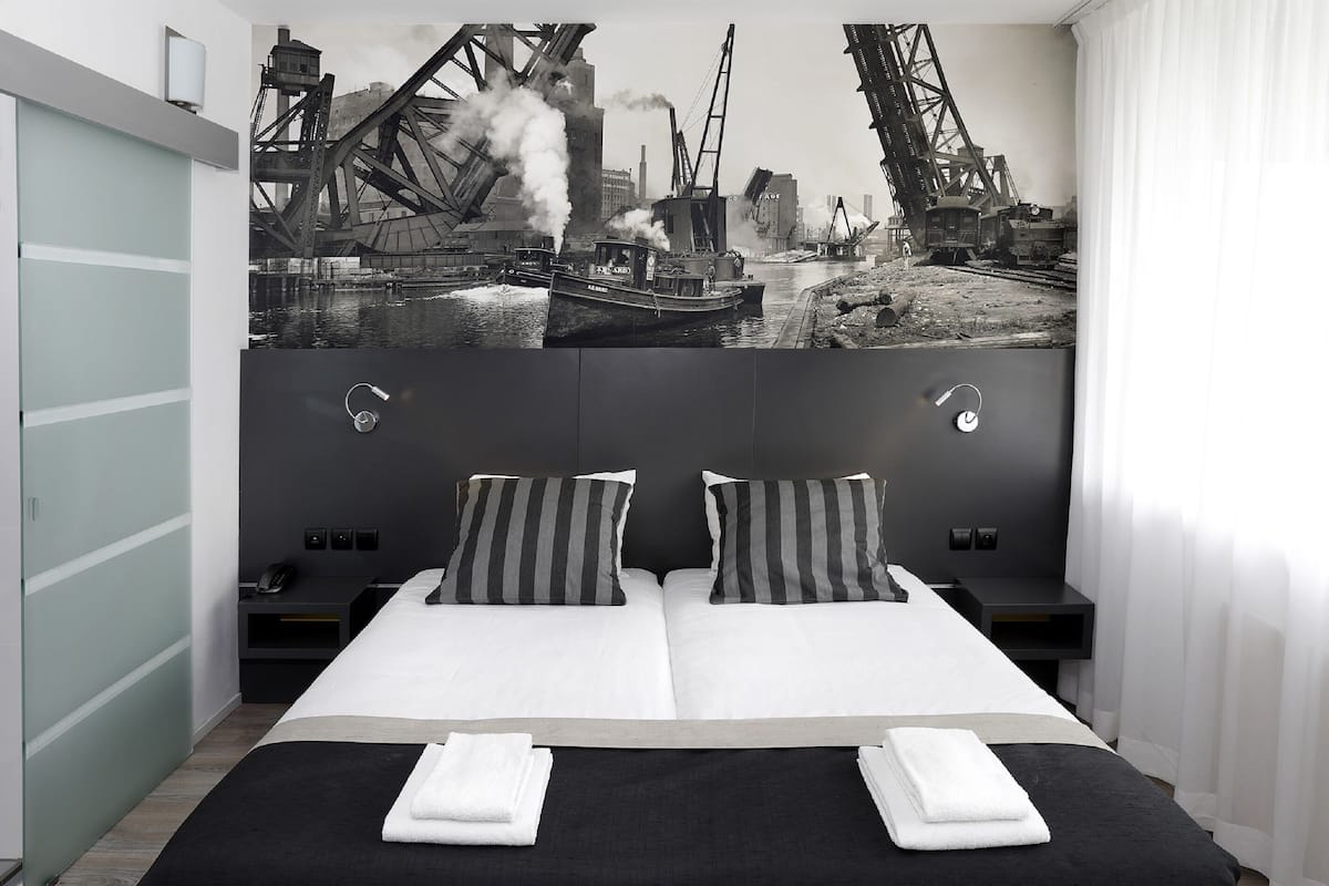 Une chambre d’hôtel moderne dotée d’un grand lit, de deux tables de chevet, d’une tête de lit ornée d’une fresque représentant un paysage urbain, et offrant une vue sur la rivière.