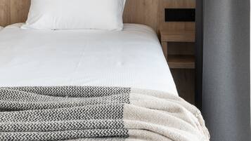 Habitación de hotel con cama, cabecera, luz de pared y vista a un atardecer sobre la ciudad.