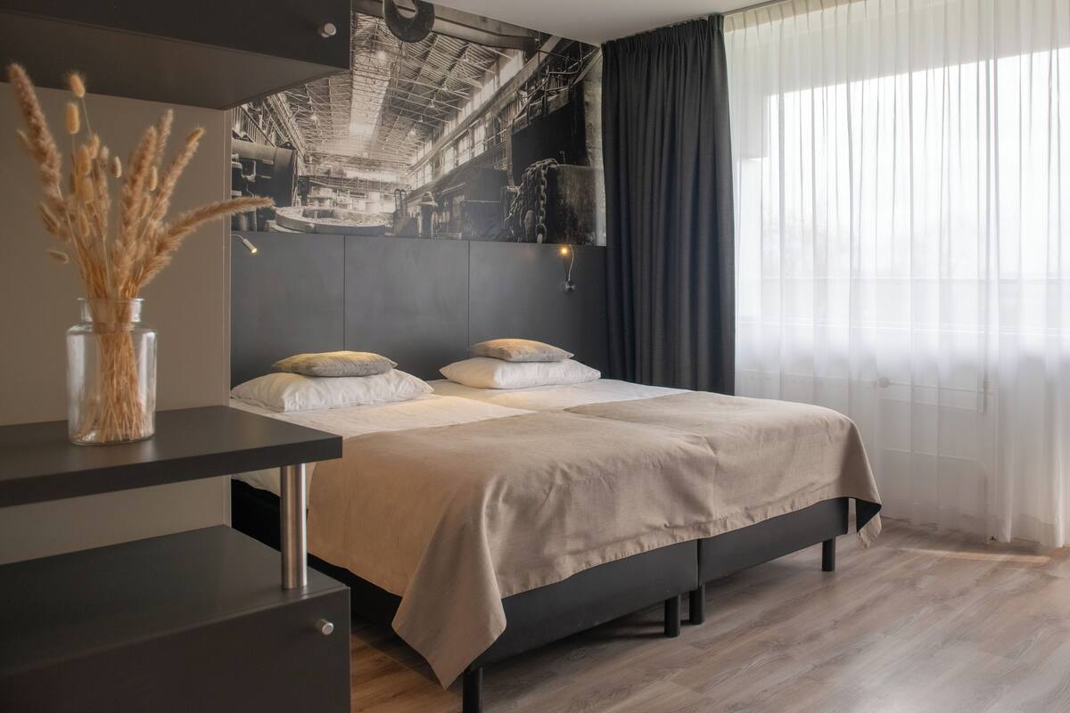 Une chambre d’hôtel moderne avec un grand lit, une table de chevet ornée d’un vase de fleurs séchées et une fresque murale.